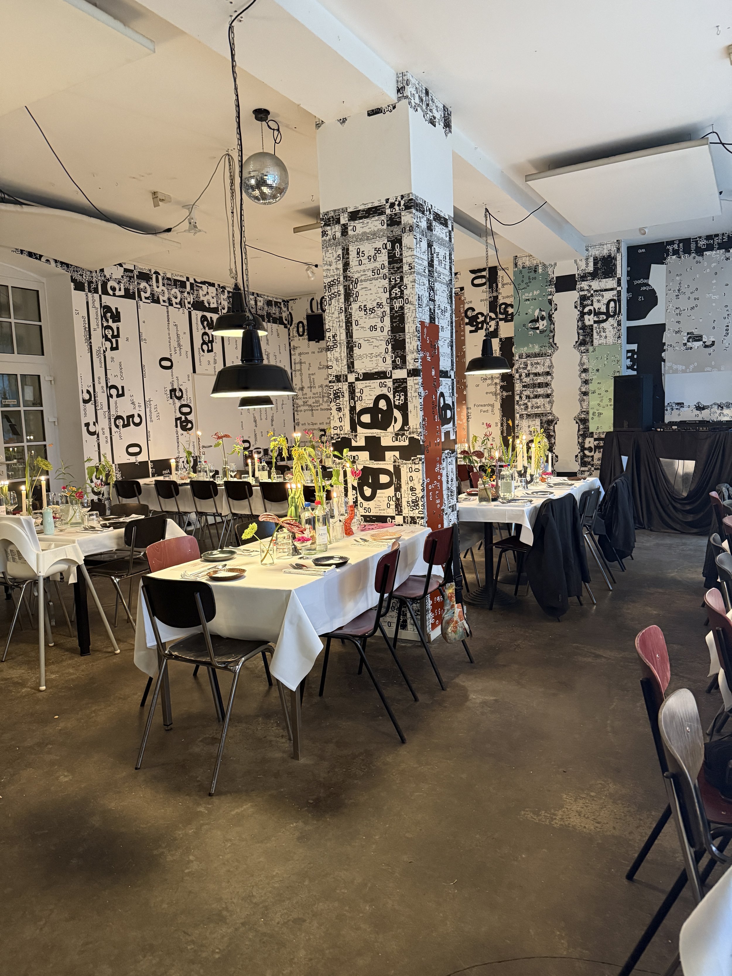 Ein Saal mit Tischen, die festlich gedeckt sind, Blumen und Kerzen, schwarze und braune Stühle, helle und dunkle Wände mit grafischen Mustern, schwarze Pendelleuchten, eine kleine Discokugel, einige Pflanzen und ein DJ-Setup im Hintergrund.
