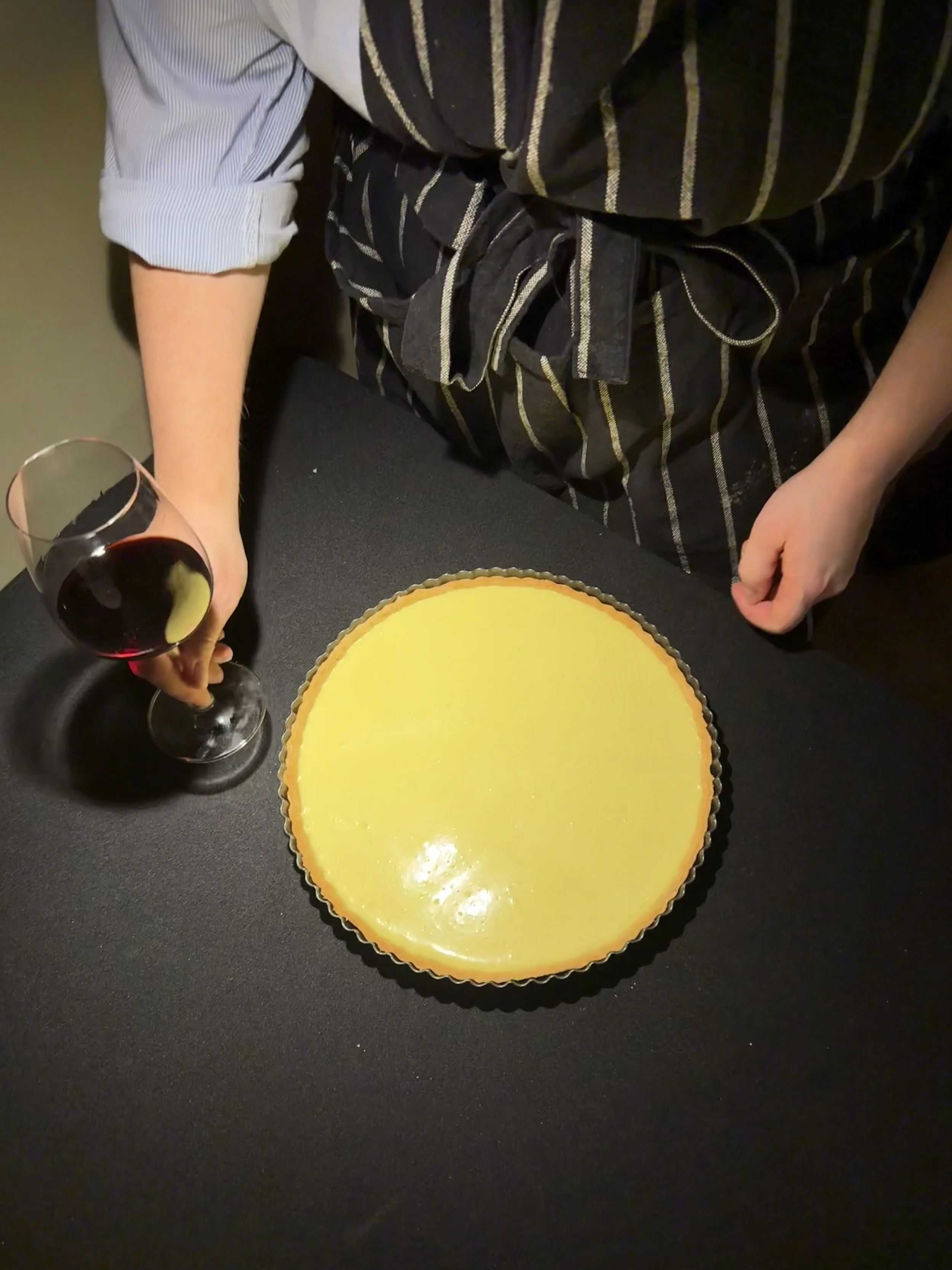 Eine Person hält ein Glas Rotwein in der linken Hand. Auf einem schwarzen Tisch liegt eine Zitronentarte. Im Hintergrund ist nur die Kleidung der Person sichtbar, die eine gestreifte Schürze trägt.