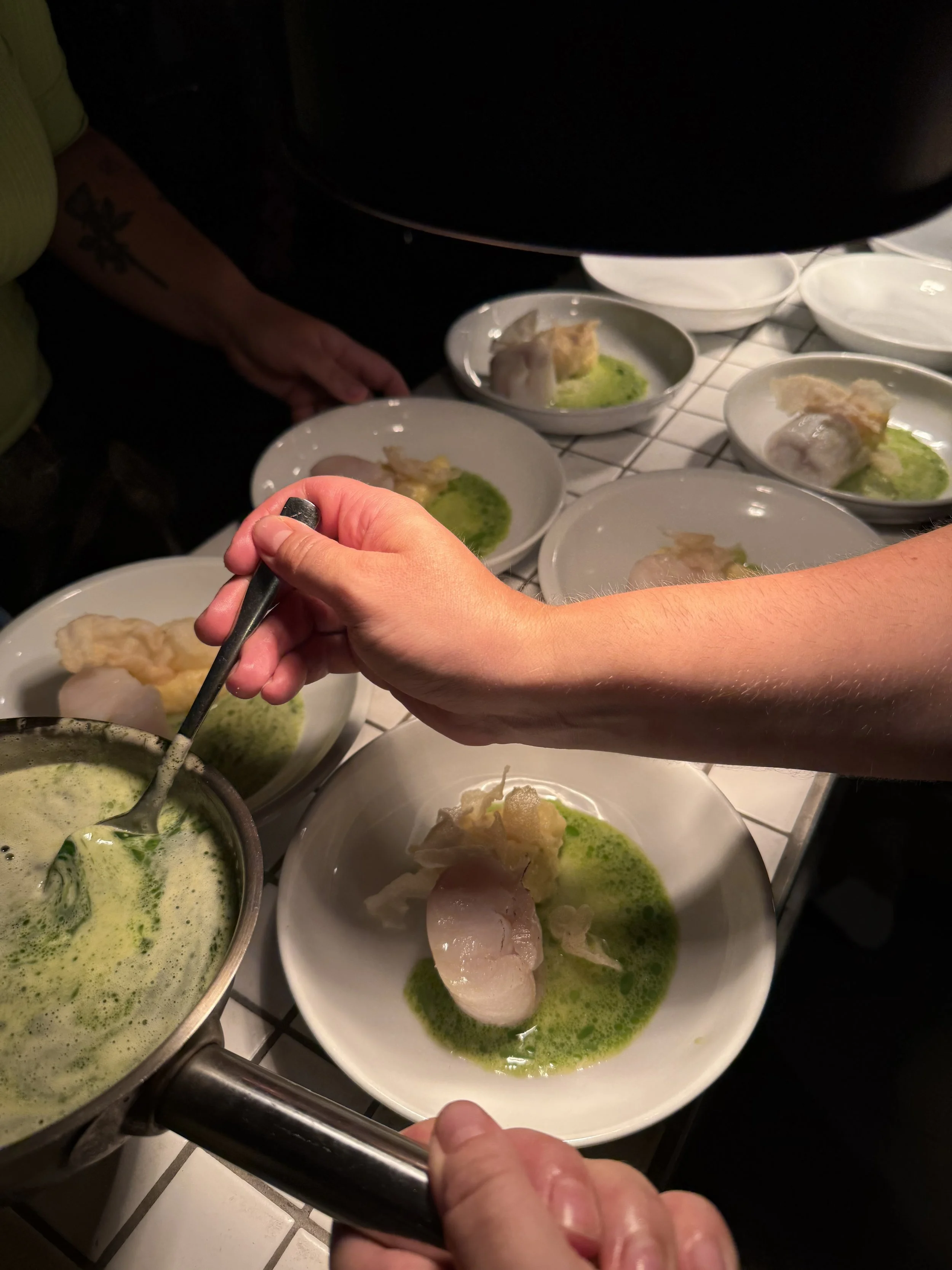 Hände, die eine grüne Soße aus einem Topf schöpft, um sie auf Teller mit weißem Fischfilet und Kartoffelbrei zu gießen. der Hintergrund ist dunkel. Der Untergrund ist eine Fliesentheke.