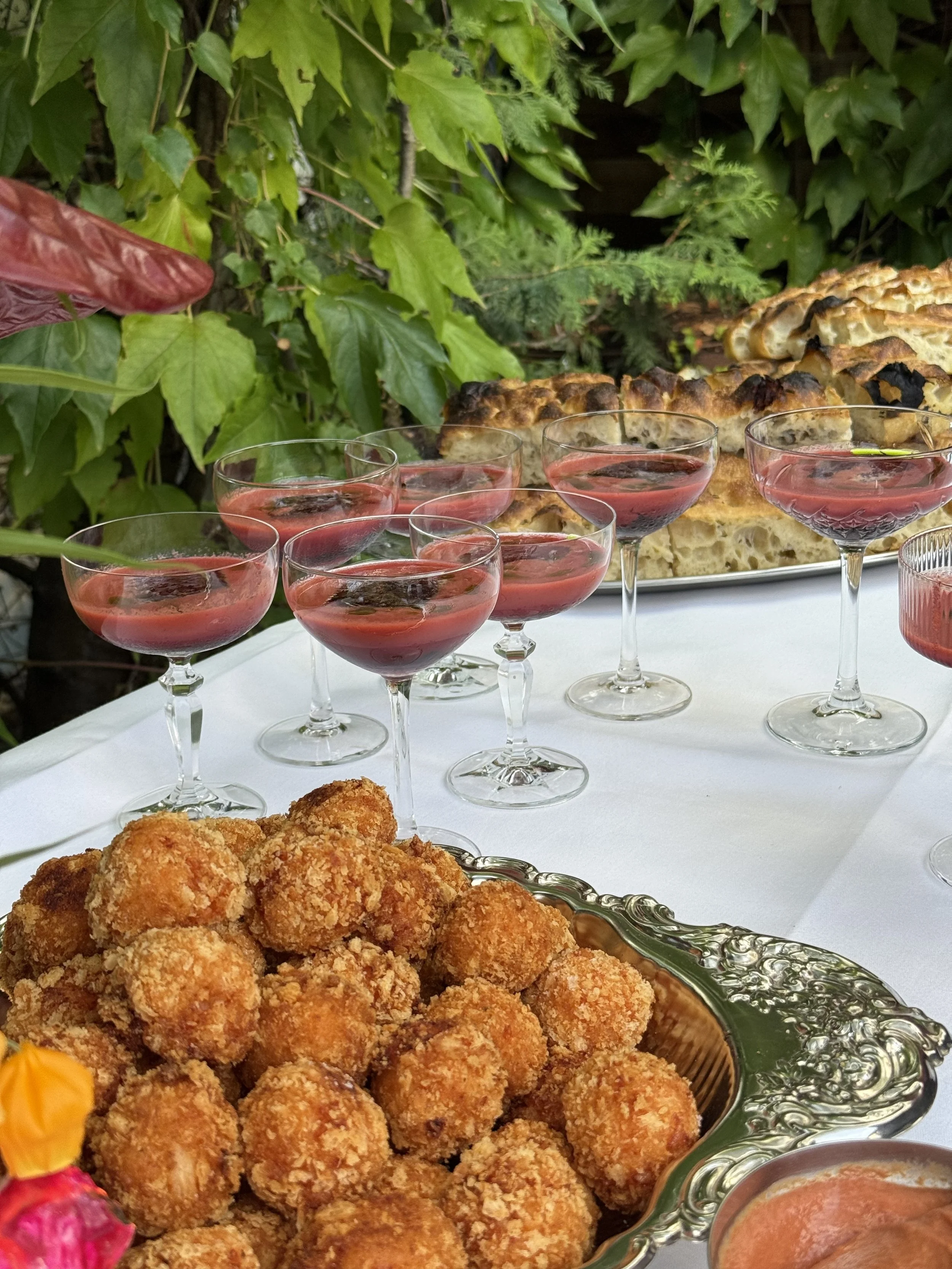 Frittierte Arancini mit roter Soße und auf einem Silbertablett, daneben ein Glas mit roter Kirschgazpacho, im Hintergrund Kräuter und Focaccia.