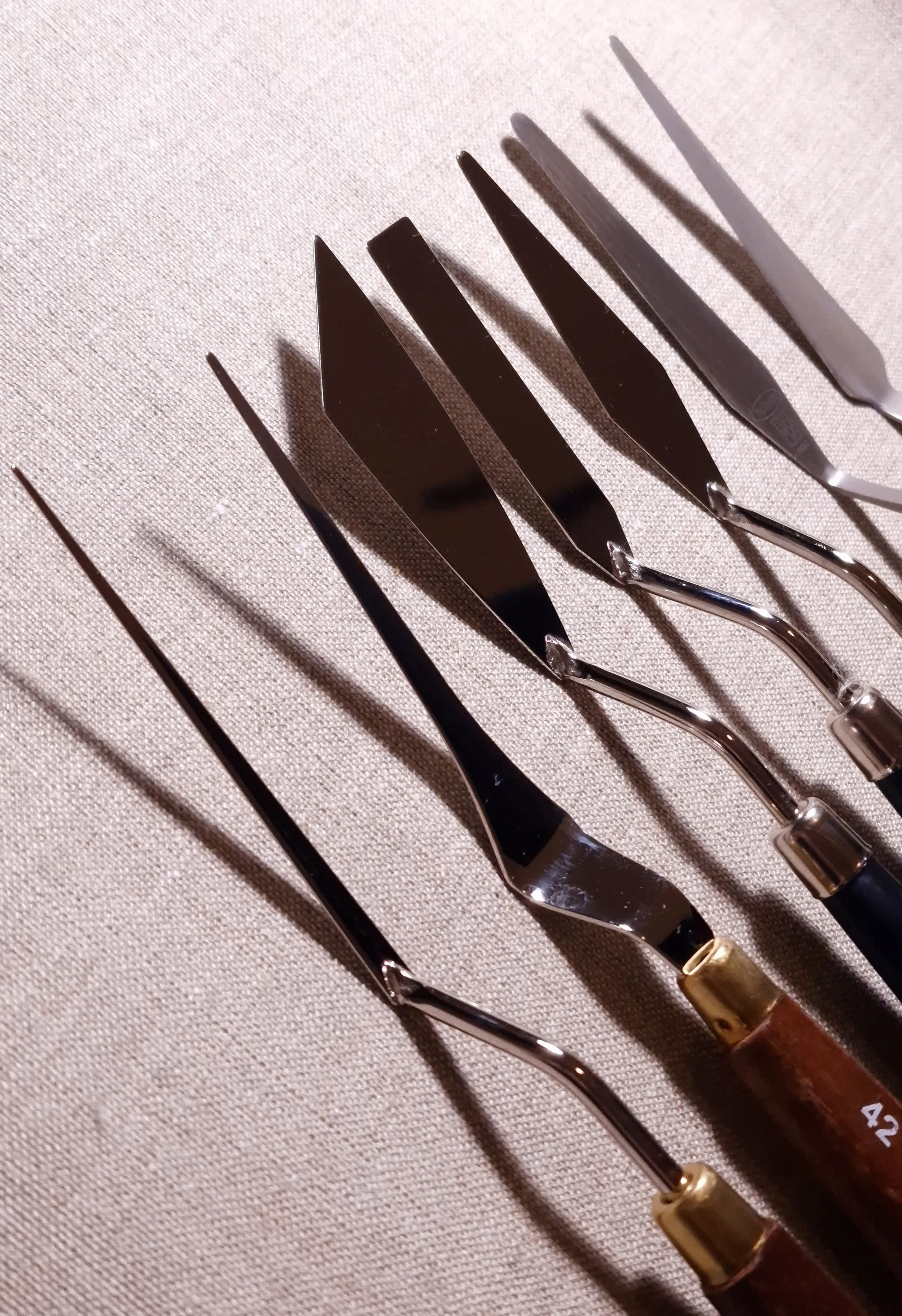 Seven painting knives with different blade shapes and geometries arranged on linen in an artist’s studio