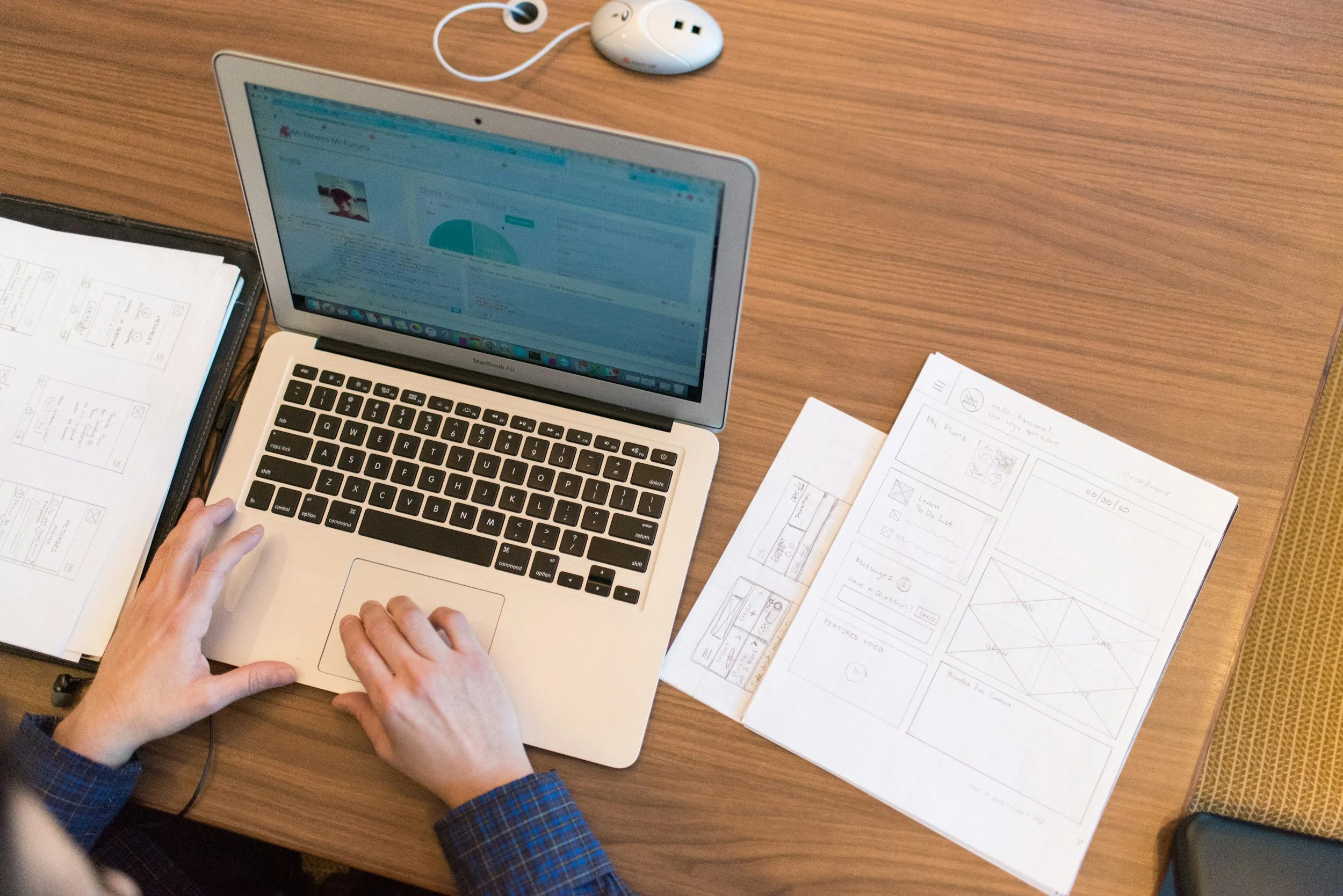 Person using a MacBook Air on a wooden table with sketches, a power strip, a charger, and papers with diagrams and notes.
