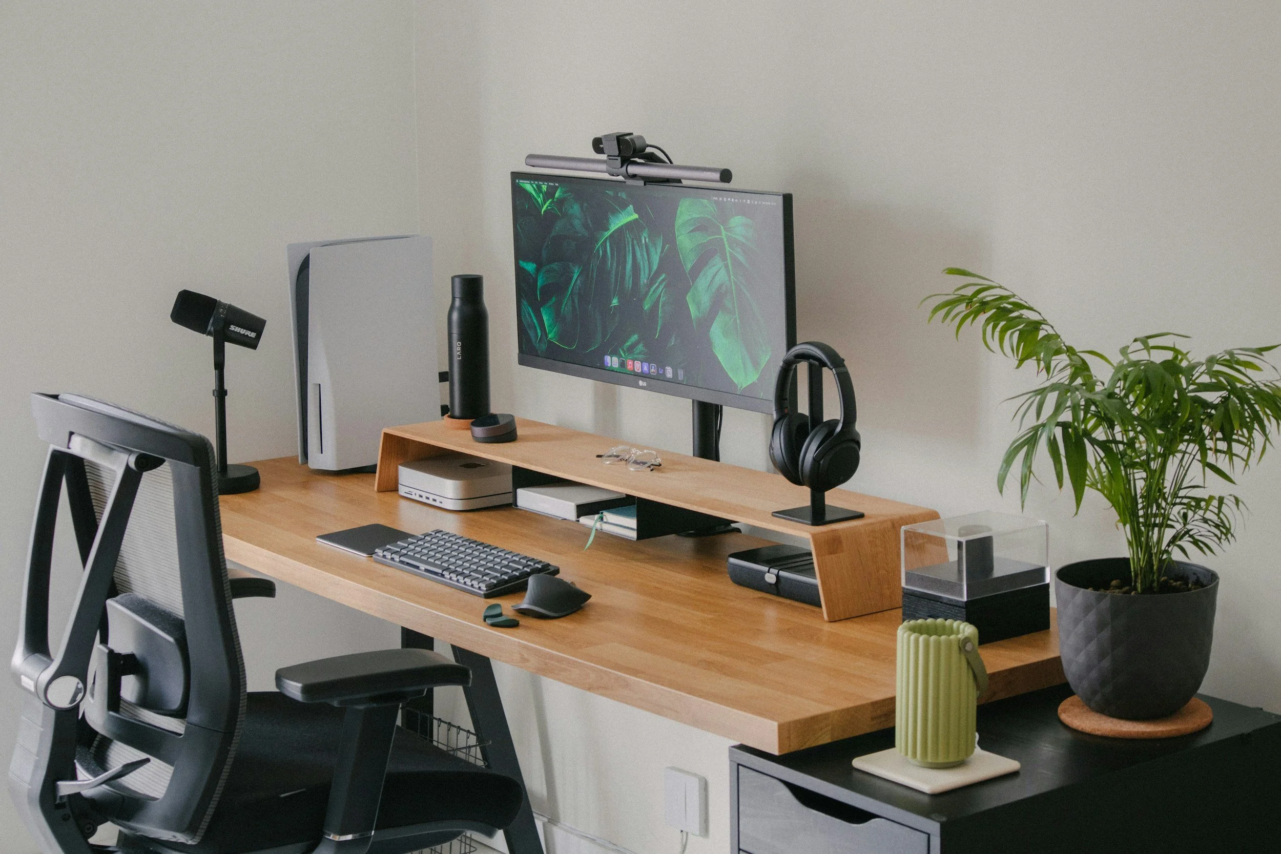A tidy home office desk setup with a computer monitor displaying green leaves, a wireless keyboard, mouse, and a pair of headphones hanging on the monitor stand. The desk also has a water bottle, a small speaker, a notebook, and some decorative items including potted plants.
