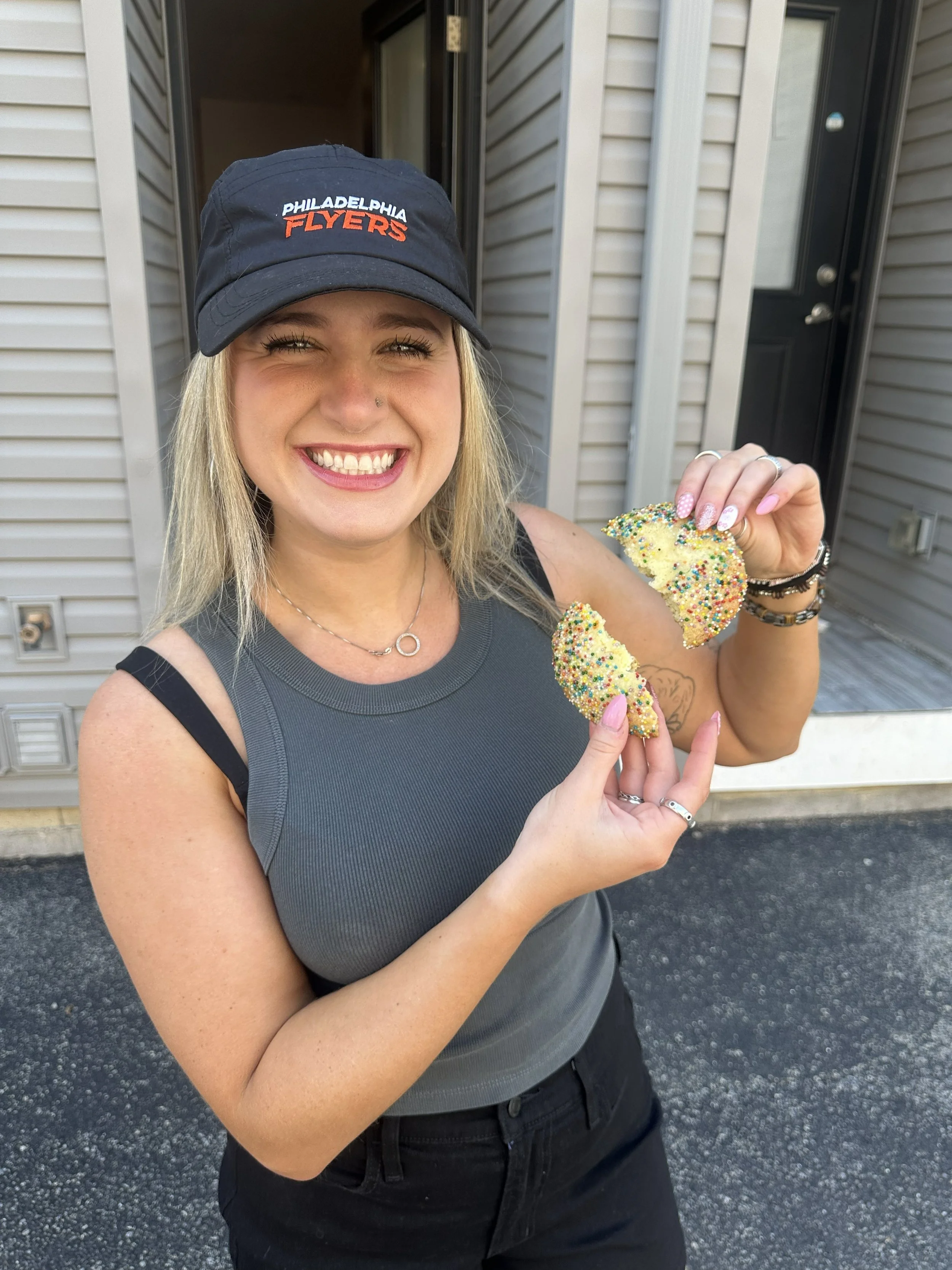 A smiling woman wearing a black Philadelphia Flyers cap and black sleeveless shirt, holding a multicolored sprinkled snack, standing outside in front of a building with siding and a black door.