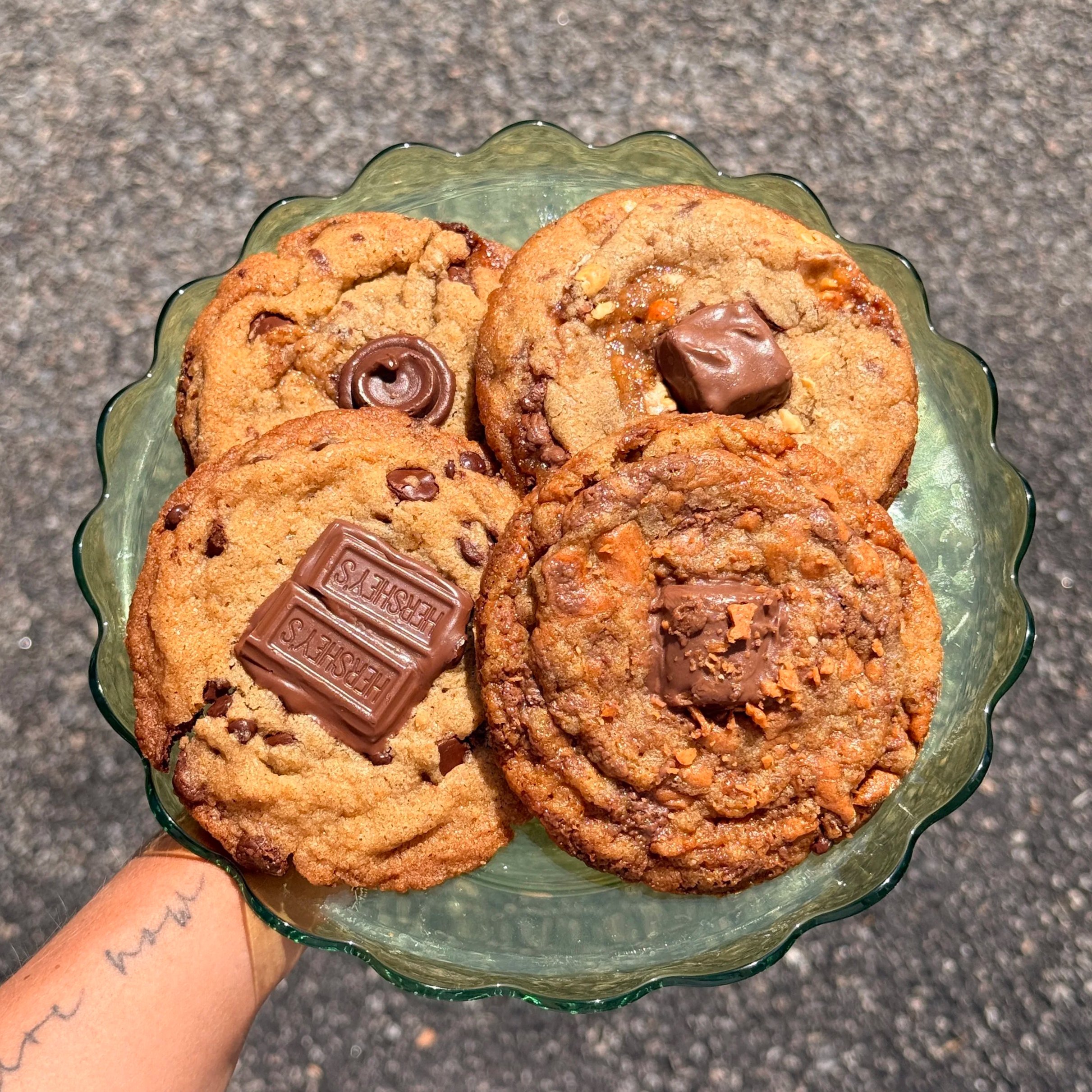 A person's hand holding a glass bowl filled with various chocolate chip cookies, some with chocolate pieces and some with nuts, against a gray textured surface background.