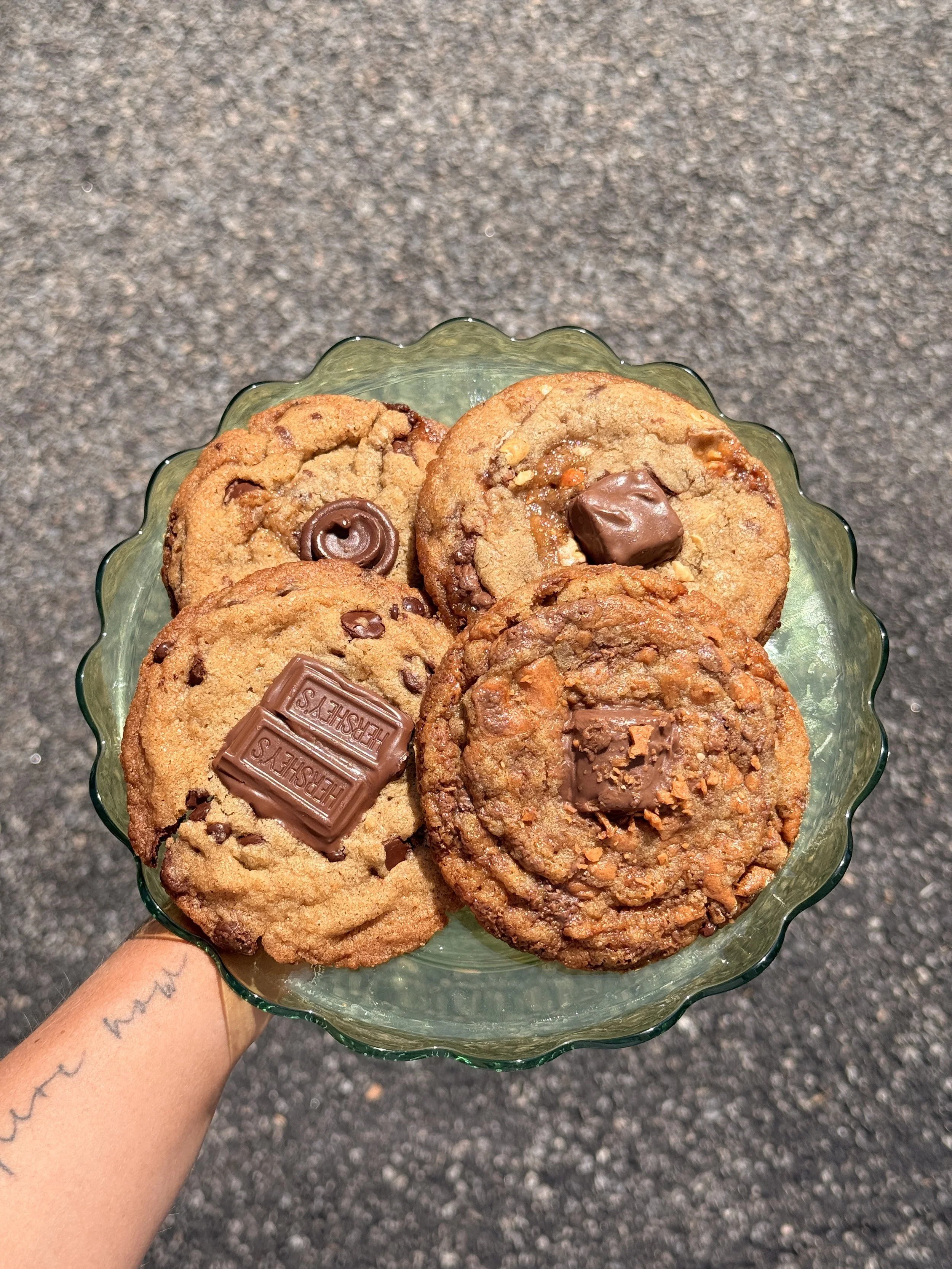 A glass dish filled with assorted cookies, including chocolate chip, peanut butter, and chocolate with Hershey's chocolates, held above a dark gray, textured surface.