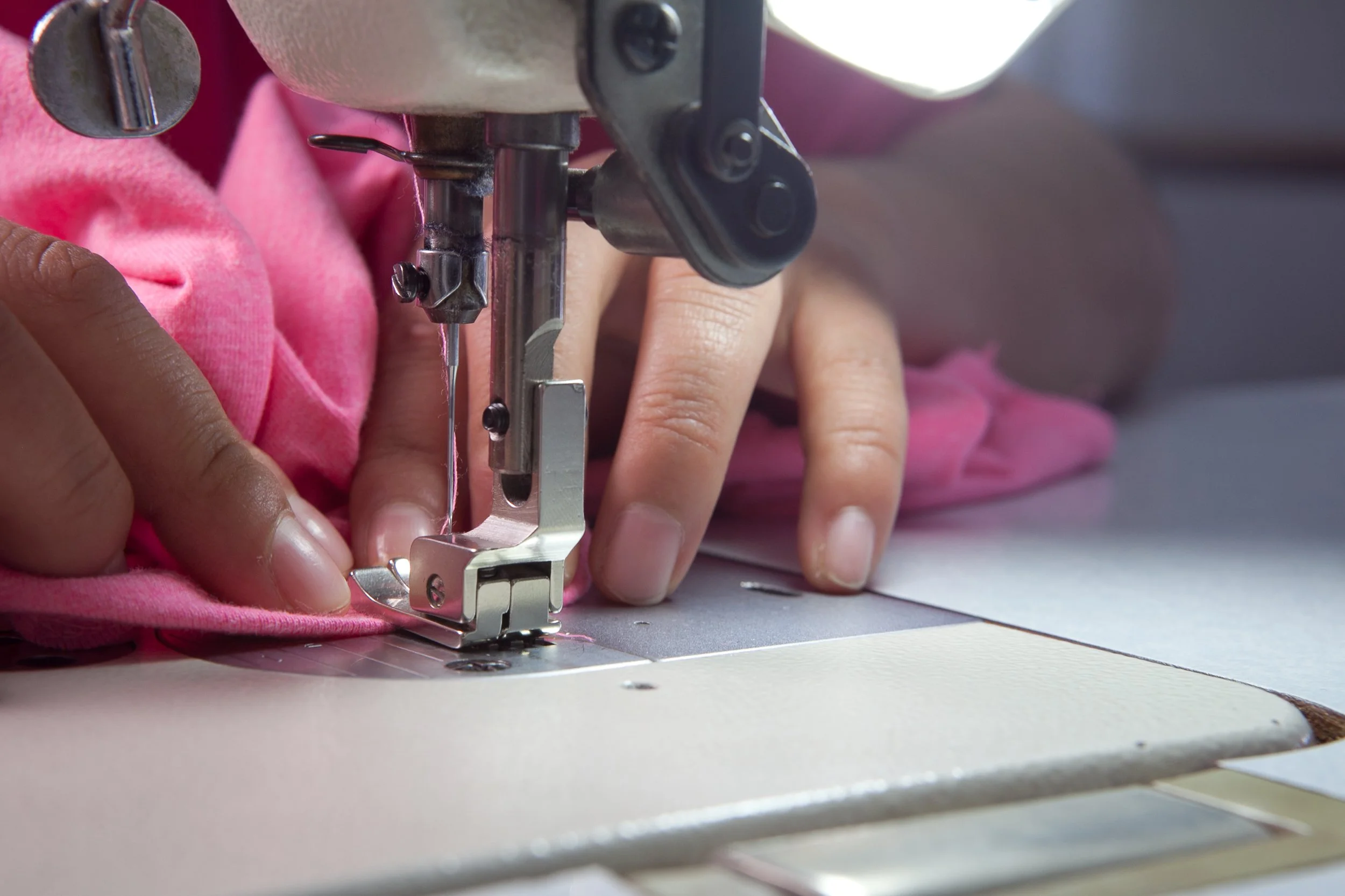 Close-up of hands guiding pink fabric through a sewing machine as it stitches fabric.