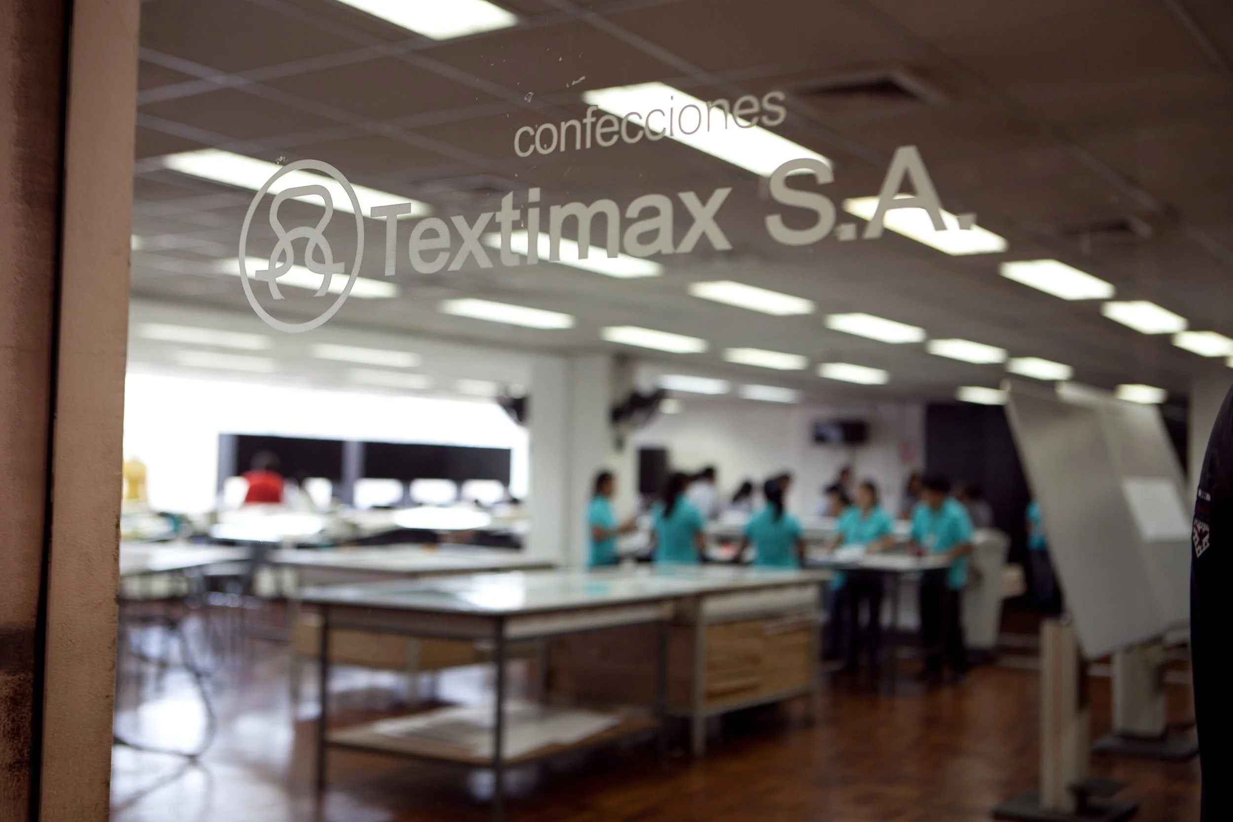 A glass window sign reads 'Textimax S.A. confecciones' with a logo of a knot. Behind the window, there is a room with multiple people in teal uniforms, working around tables in a brightly lit space with a drop ceiling.