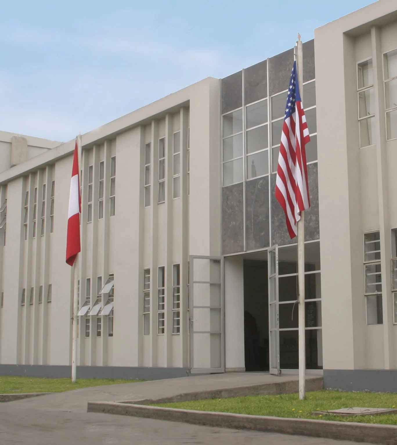 Modern white building with multiple windows and two flags, the United States flag and another flag, flying on poles outside.