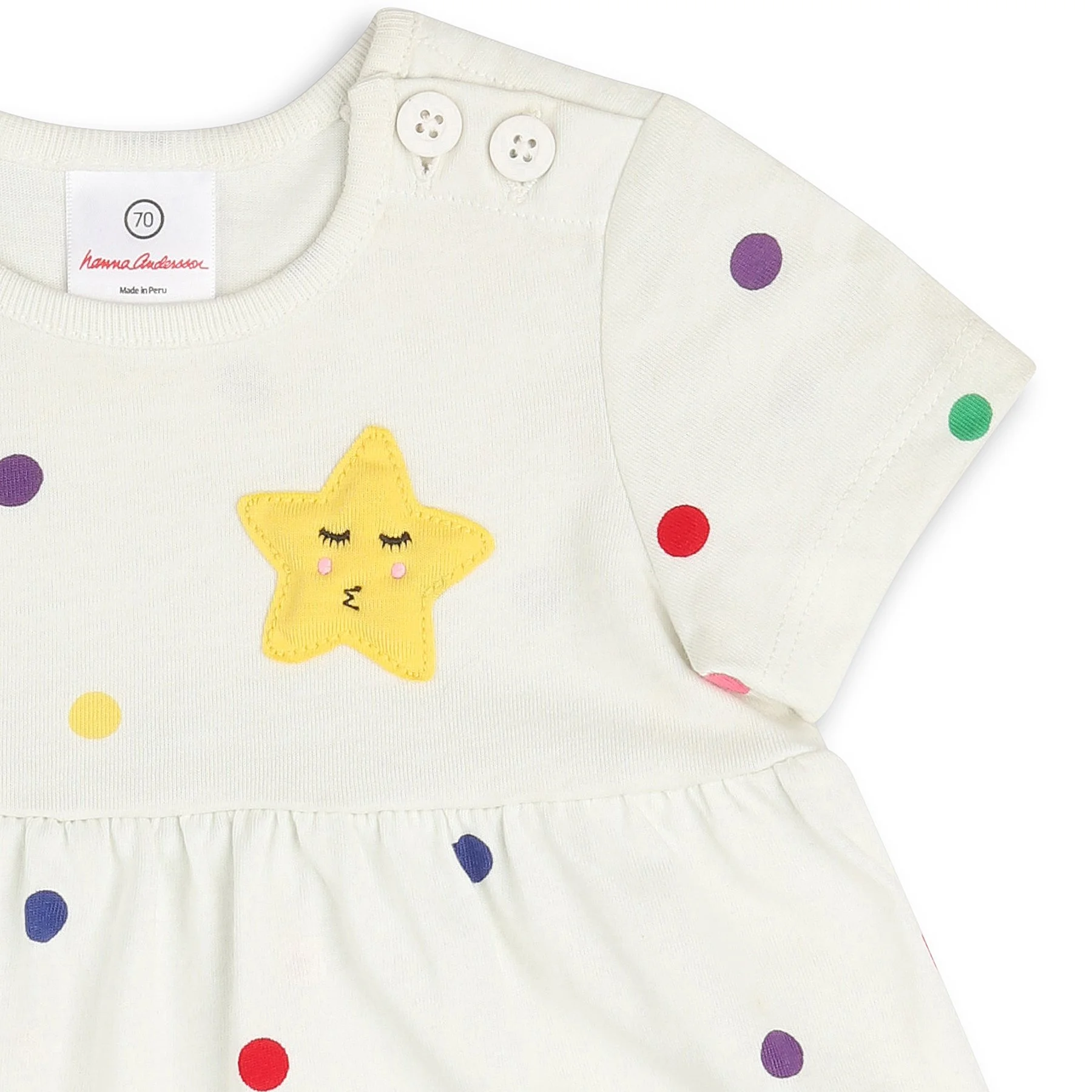 Close-up view of a cream-colored children's dress with colorful polka dots and a smiling yellow star with closed eyes embroidery on the front. The dress has two white buttons on the shoulder and a label indicating size 70 and made in Peru.