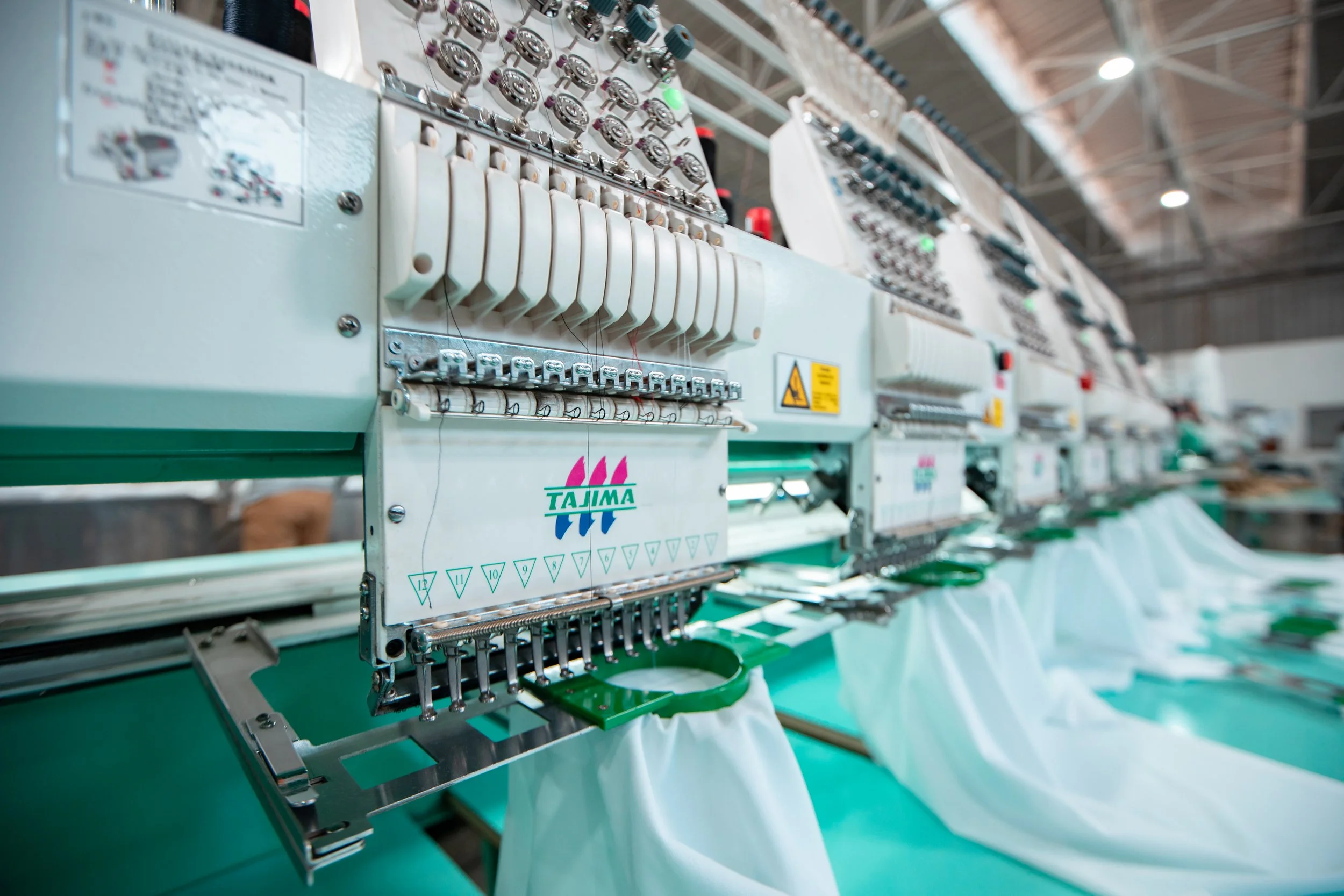 Close-up view of an industrial embroidery machine in a factory with multiple machines in the background. The machine has a metal frame with various buttons, dials, and threading mechanisms, and a green surface underneath.