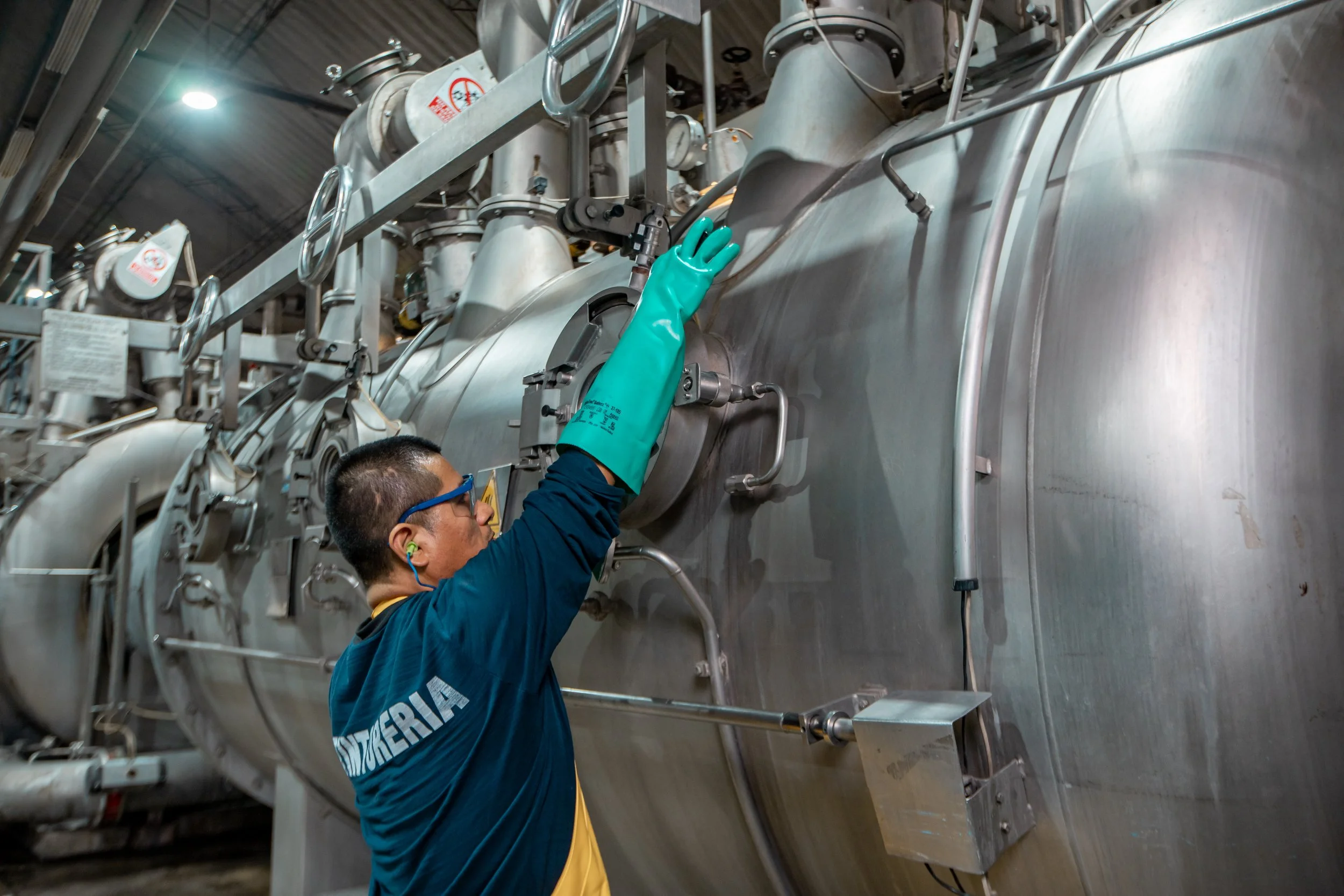 A worker wearing teal protective gloves, safety glasses, and a dark blue uniform with yellow accents is adjusting a large industrial machine made of metal, with numerous pipes and gauges, in an industrial setting.