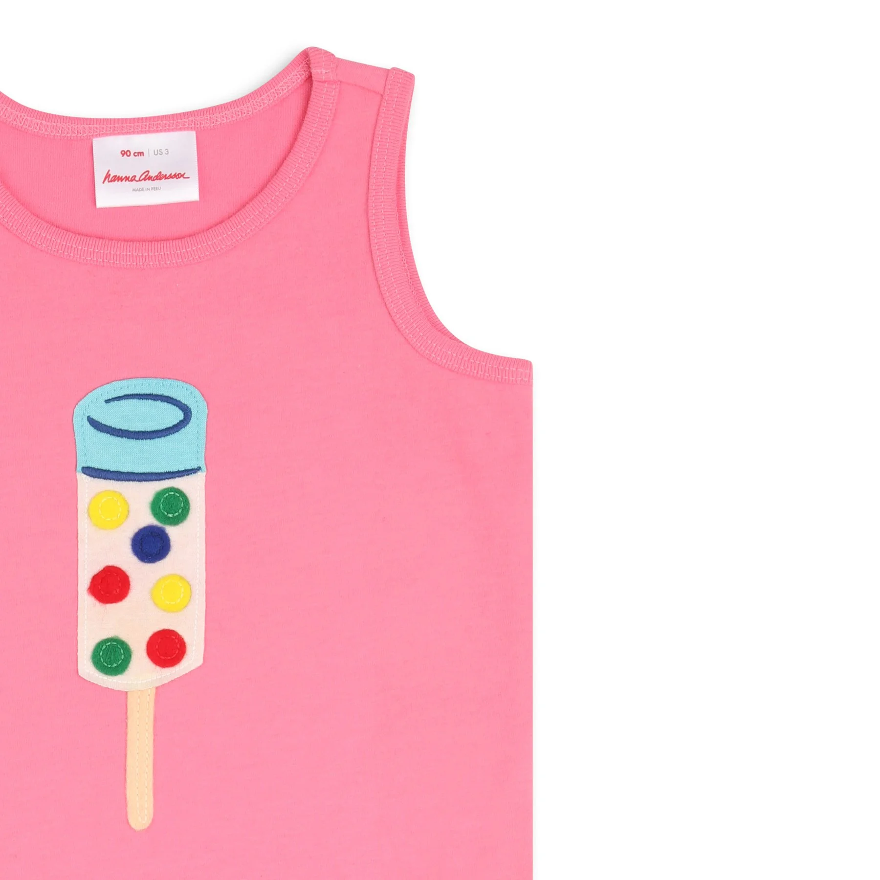 A pink sleeveless shirt with a graphic resembling a colorful toy capsule on the front, and a label indicating the size 90 cm US 3 by Hanna Andersson, made in Peru.