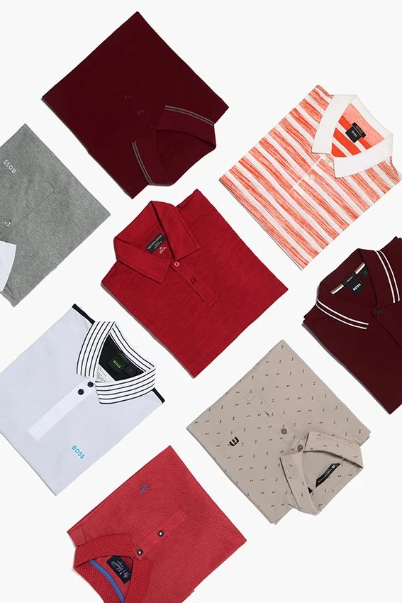Array of folded men's polo shirts in various colors and patterns, including red, white with black stripes, beige with small black dots, and solid maroon, arranged on a white background.