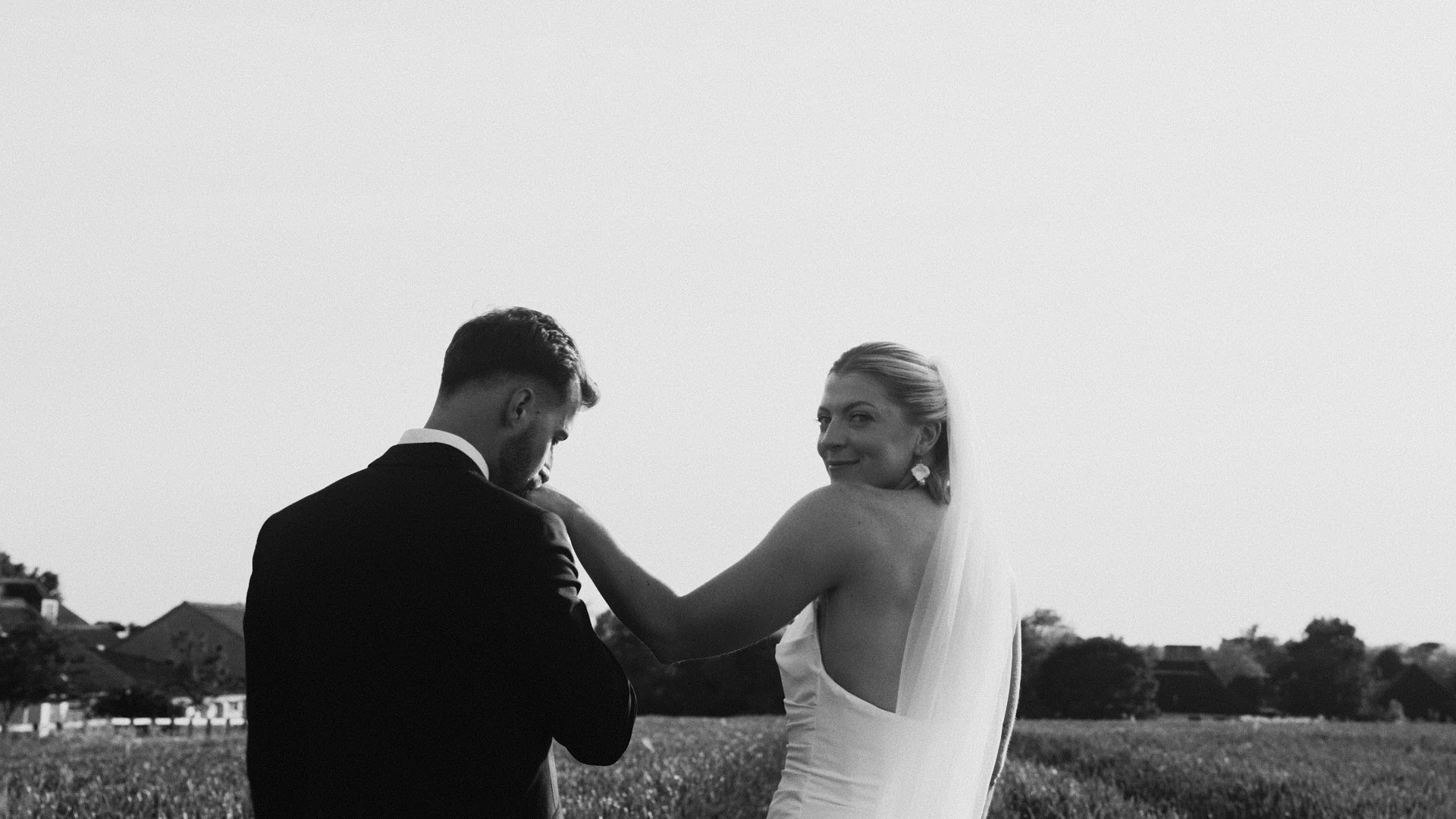 A black and white photo of a bride and groom standing outdoors, with a few houses and trees in the background, during a wedding ceremony.