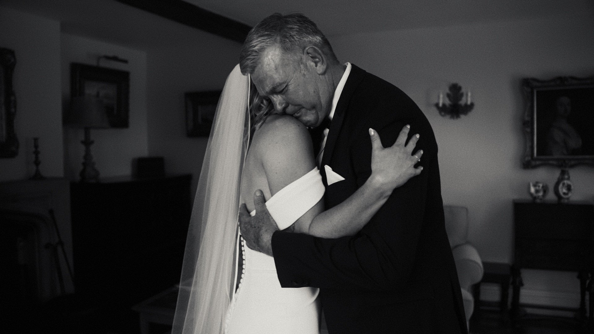 A black and white photo of a man in a tuxedo and a woman in a wedding dress sharing an embrace, likely during a wedding or special event, inside a decorated room.