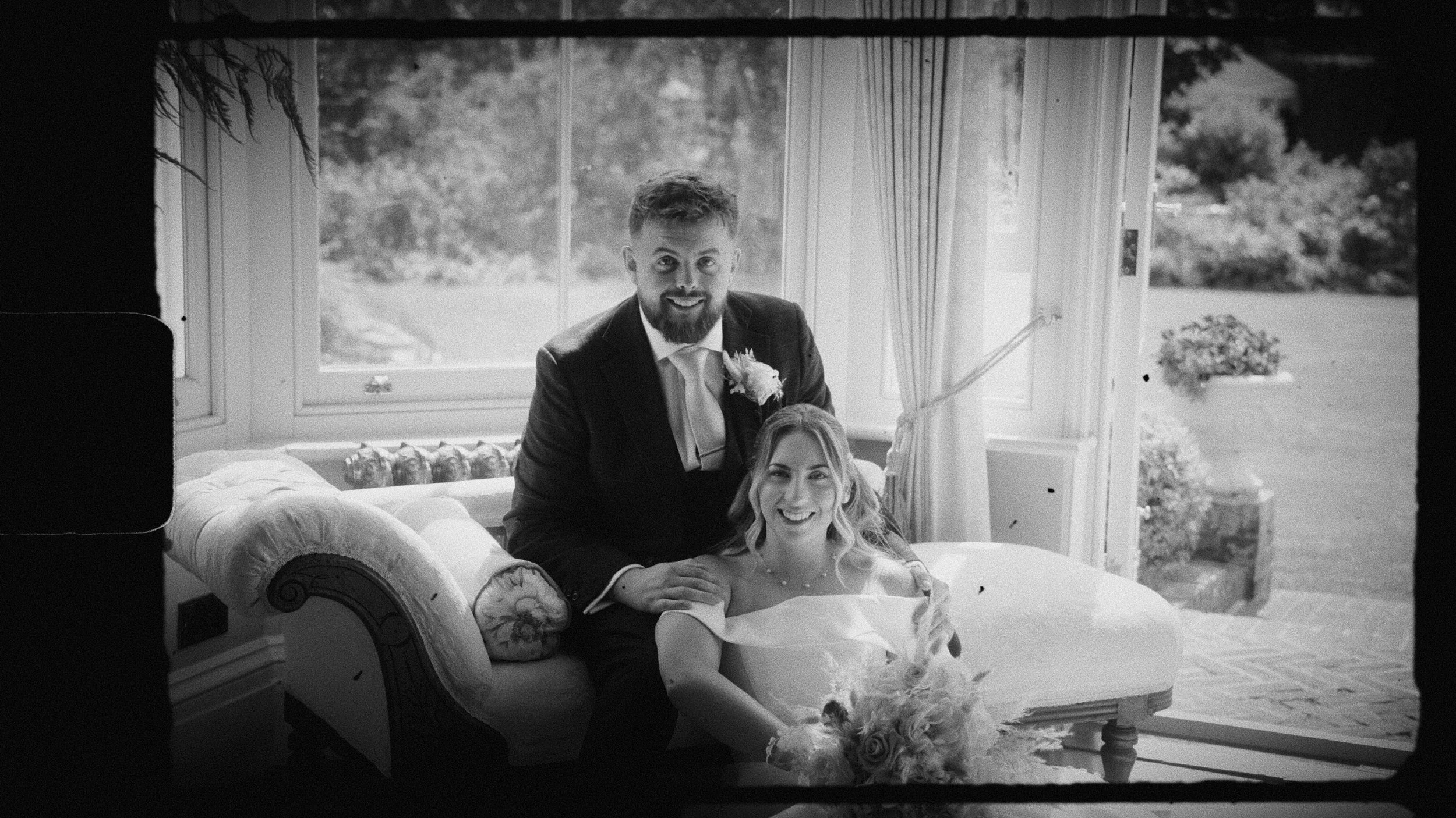 A smiling bride and groom indoors, sitting on a vintage sofa near a large window, with garden scenery outside.