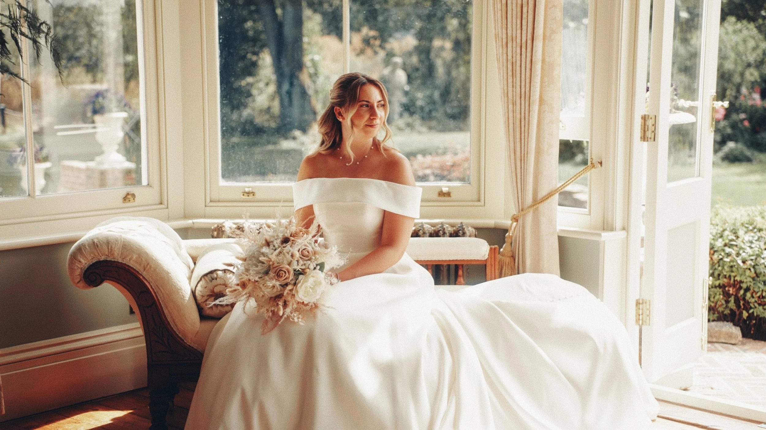 A woman in a white wedding dress sitting on a vintage sofa near large windows with a view of greenery outside, holding a bouquet of pale roses and other flowers.