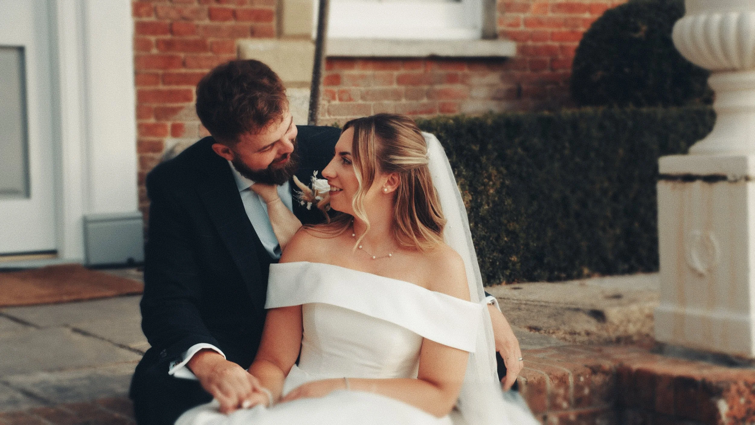 A couple dressed in wedding attire sitting close together outside a brick building, sharing a joyful moment and smiling at each other.