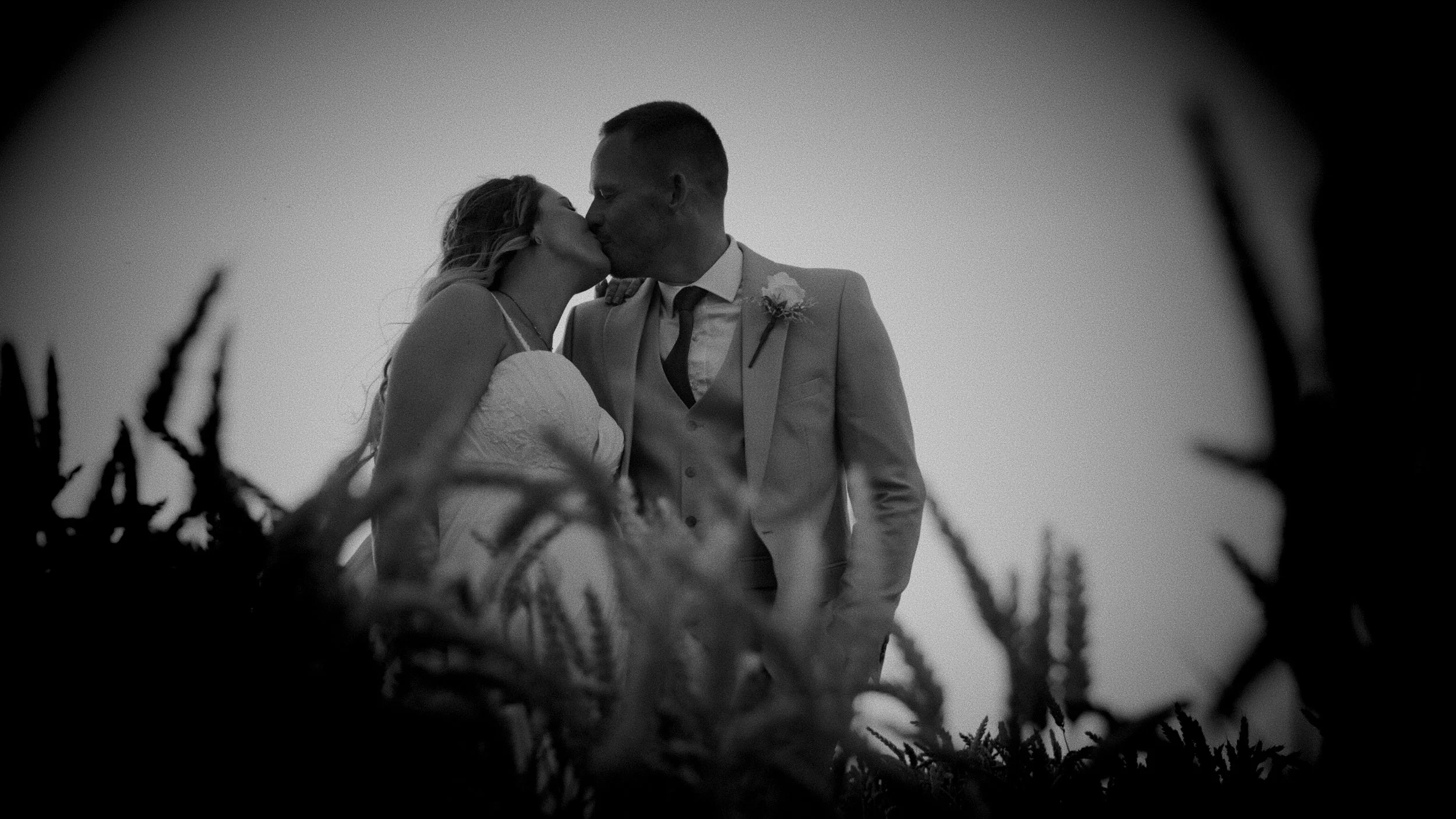 A black-and-white photograph of a couple kissing, dressed in wedding attire, outdoors with a clear sky, framed by plants or branches in the foreground.
