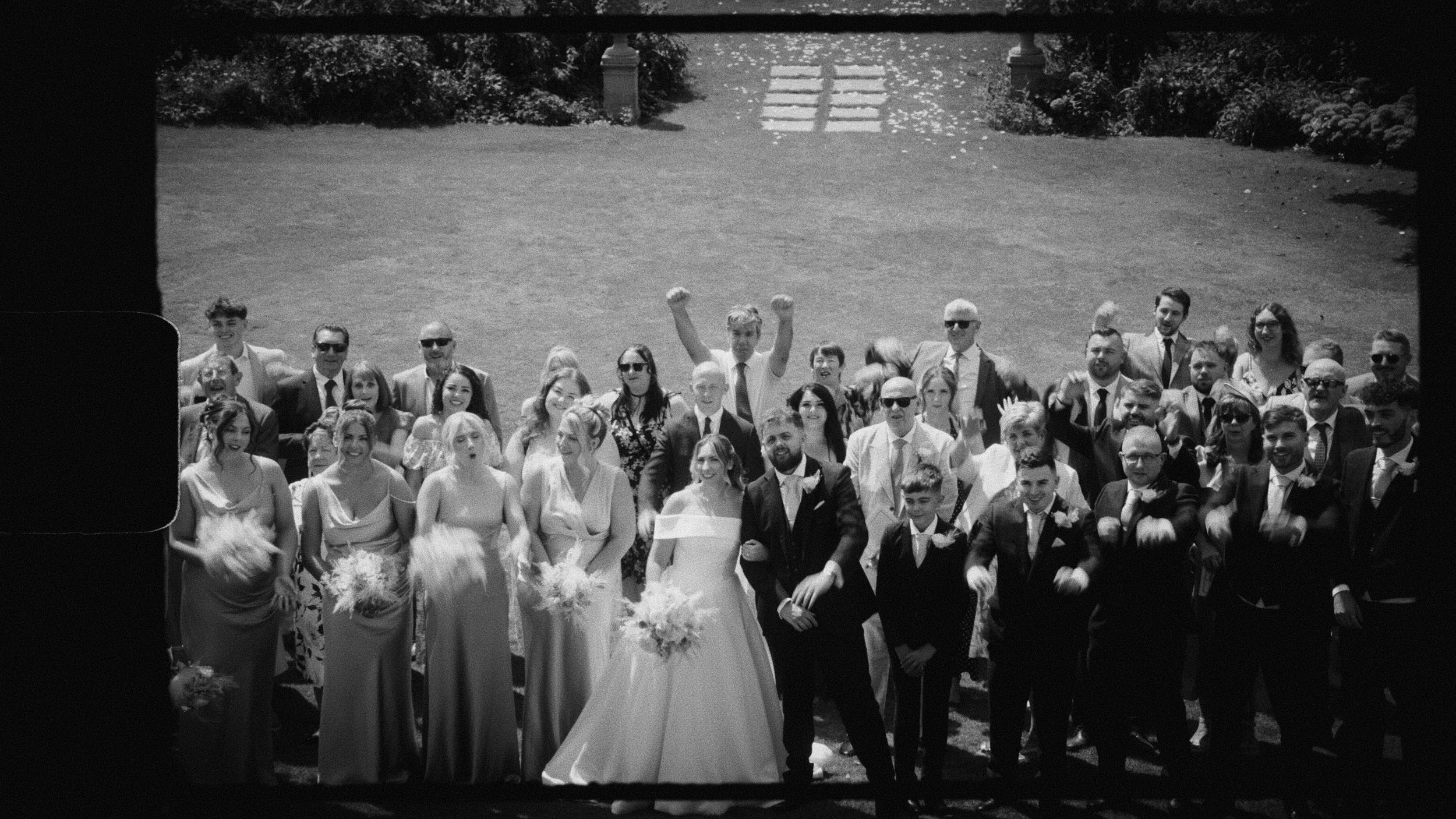 A black and white photo of a large group of people, including men, women, and children, dressed in formal attire, gathered outdoors on a lawn. Some individuals are raising their arms or waving, and the group appears to be celebrating or attending a special event, possibly a wedding.