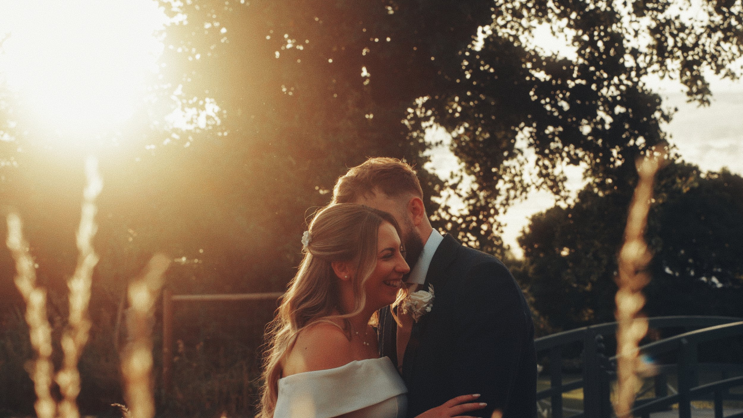A bride and groom standing close together outdoors during sunset, smiling and sharing an intimate moment, surrounded by blurred trees and a fountain in the background.