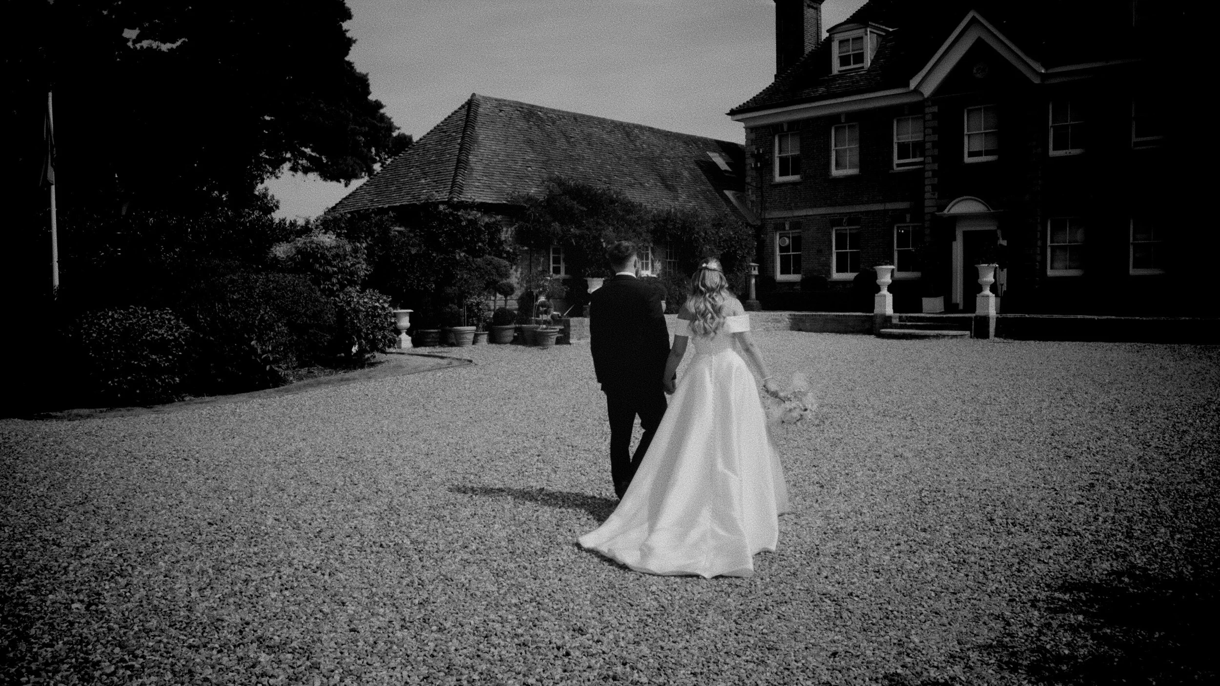 A bride and groom walking hand in hand outside of a large brick house, with the bride wearing a white wedding gown and holding a bouquet, in black and white photograph.