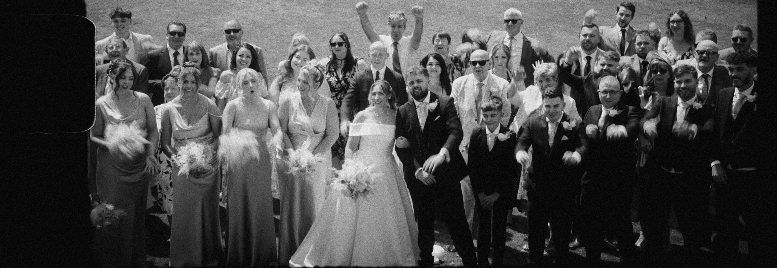 Black and white photo of a large group of wedding guests outdoors. The bride and groom are in the center, smiling, with the bride holding a bouquet. Guests are dressed in formal attire, some wearing sunglasses, many smiling or cheering, some making celebratory gestures.