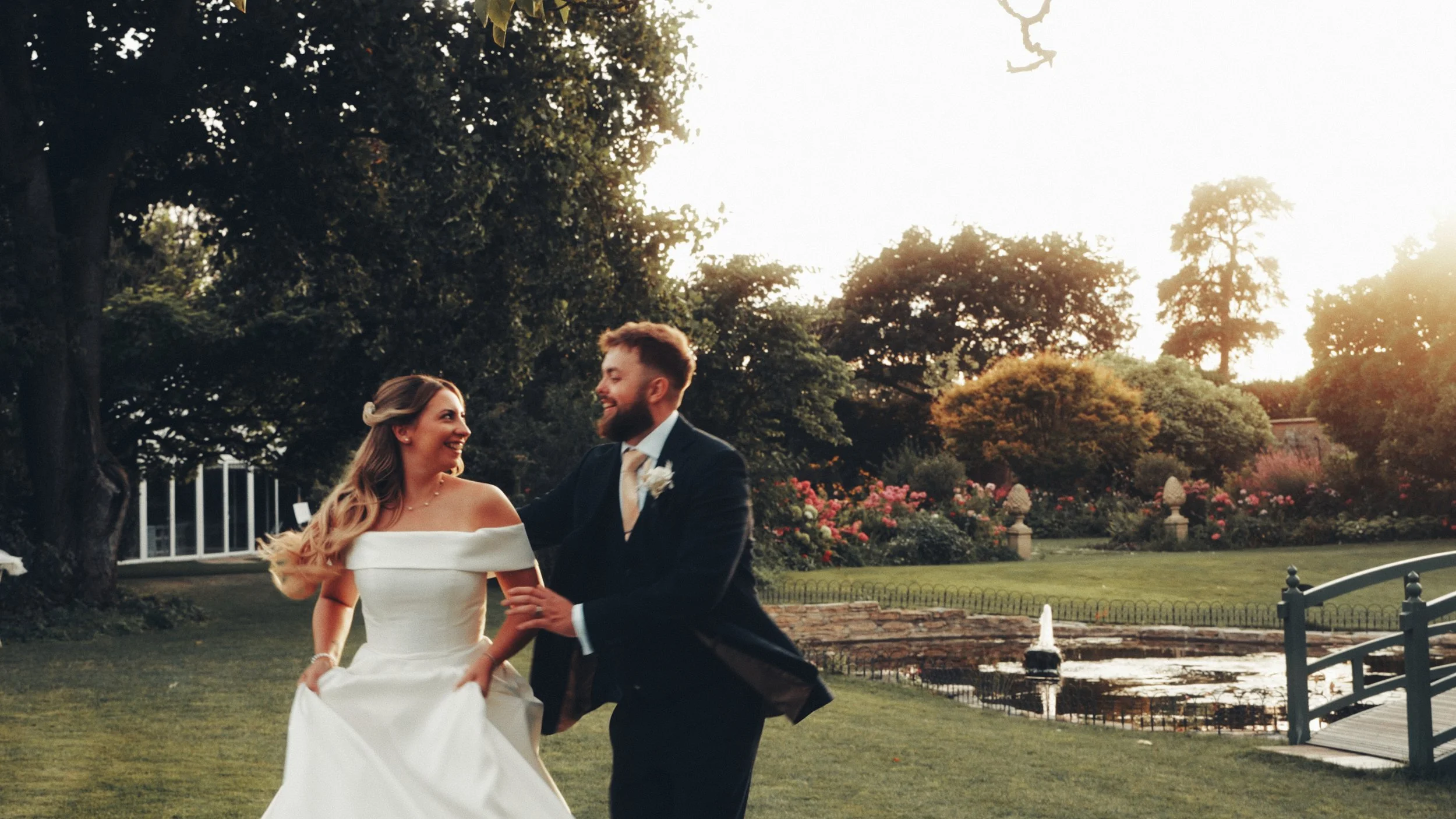 A bride and groom dancing outdoors in a garden during sunset, surrounded by trees, flowers, and a fountain.