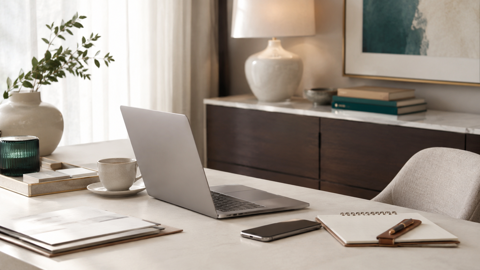 A modern home office desk with a laptop, smartphone, notebooks, pens, a cup and saucer, and a decorative plant in a white vase. In the background, a sideboard with books, a large ceramic vase, and framed artwork.