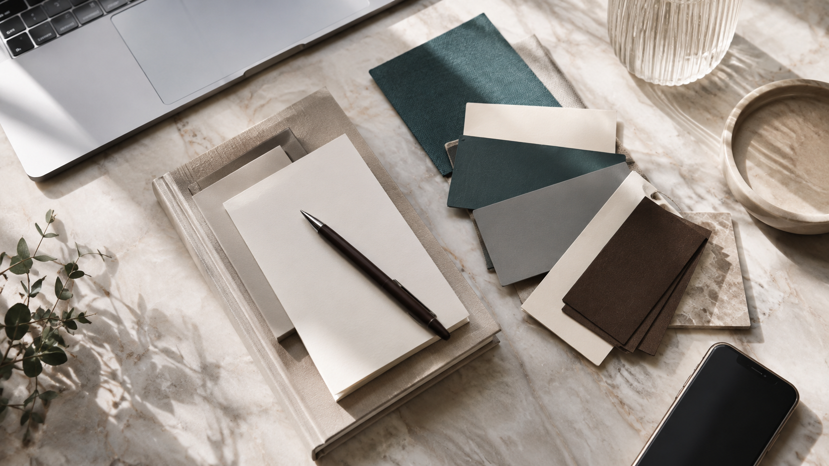 Design workspace with fabric swatches, a notebook, pen, laptop, plant, and stone bowls on a marble surface.