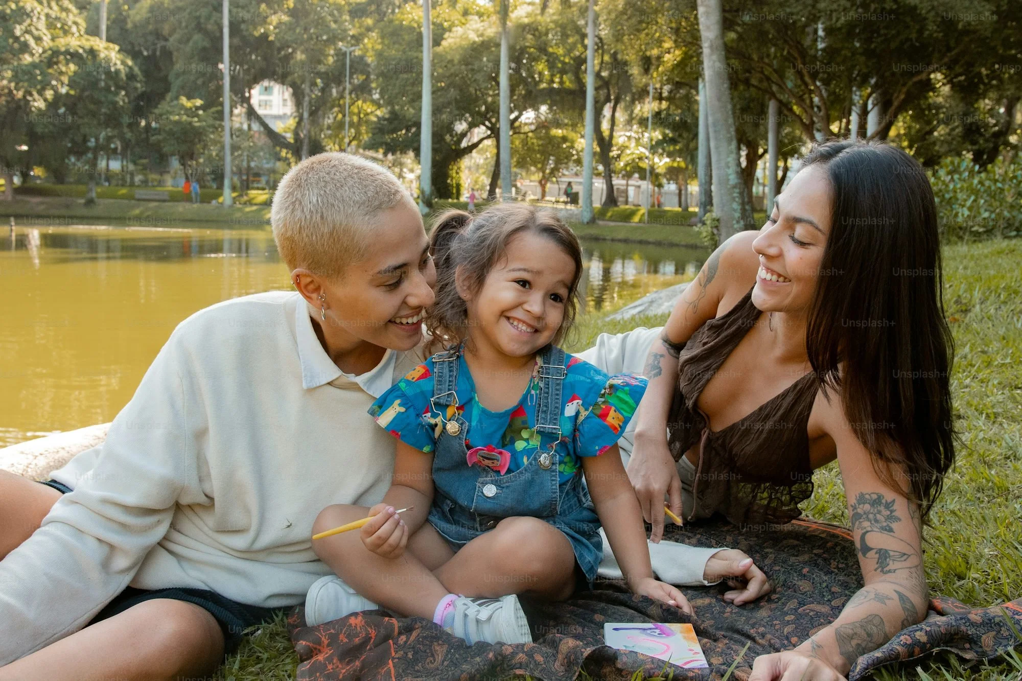 Three women and a young girl sitting by a pond in a park, smiling and enjoying each other's company on a sunny day.