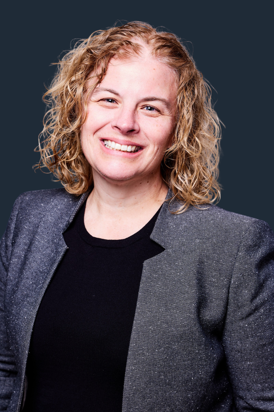 A woman with curly blonde hair smiling, wearing a dark blazer and black top, against a dark background.