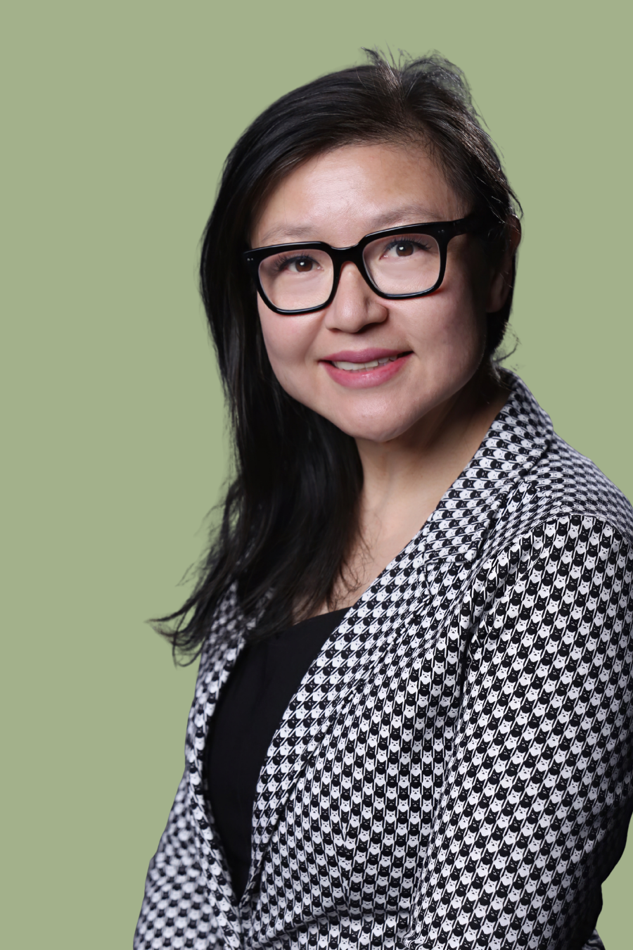 Portrait of a woman with black hair and glasses, smiling, wearing a patterned blazer over a black top, against a green background.