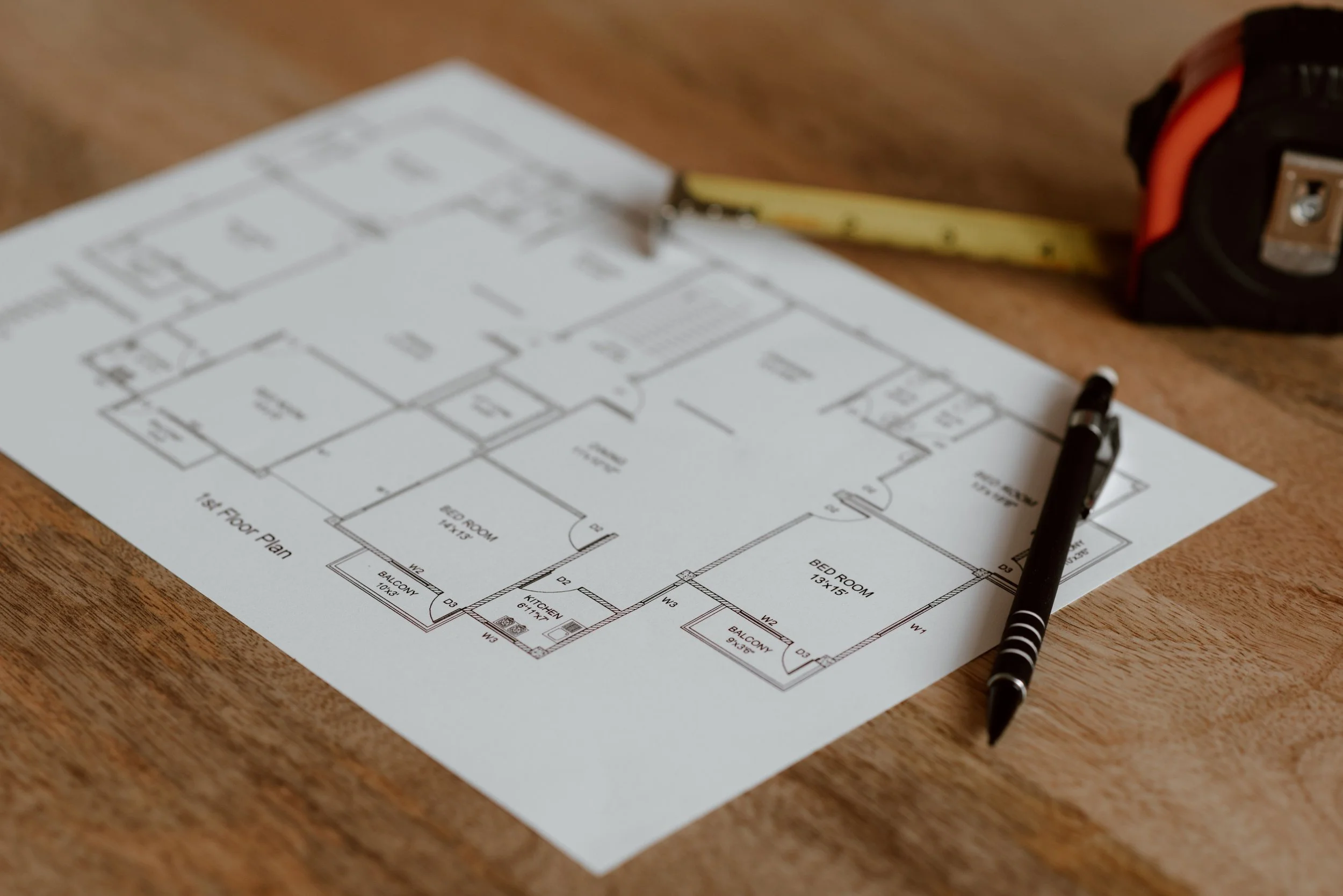 Architectural blueprint on a wooden surface with a black pen, measuring tape, and a black and red tape measure nearby.