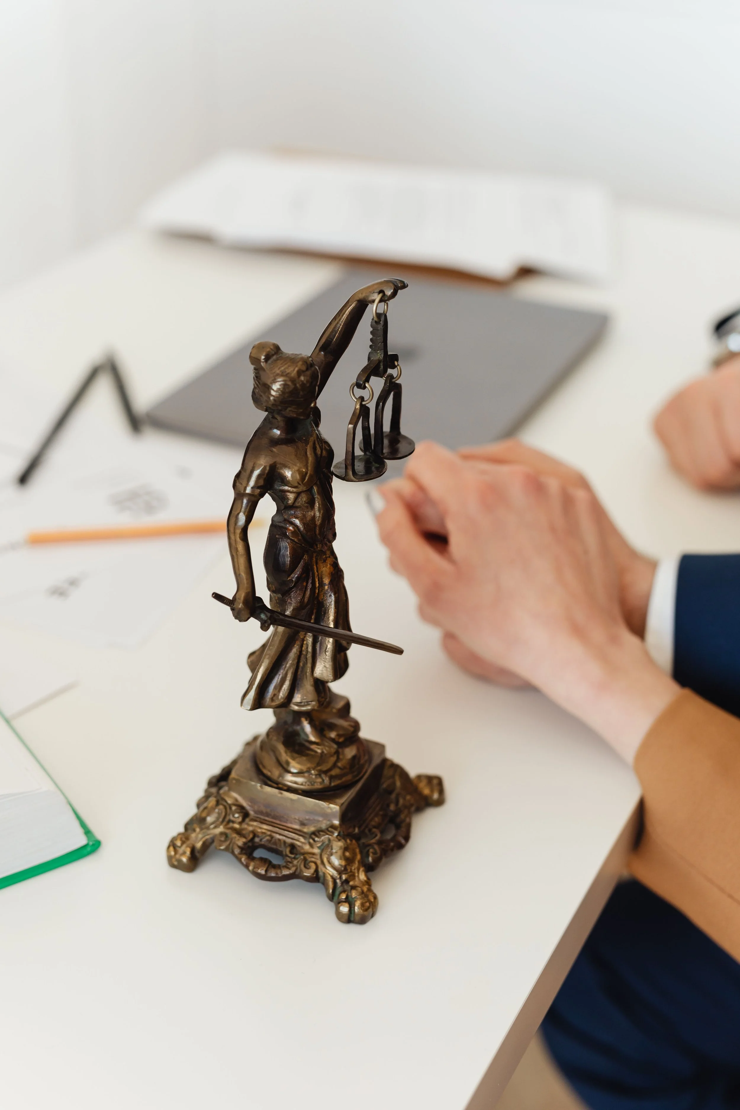 A bronze statue of Lady Justice holding scales in her left hand and a sword in her right hand, placed on a table with a person wearing a suit and glasses, and various office supplies in the background.
