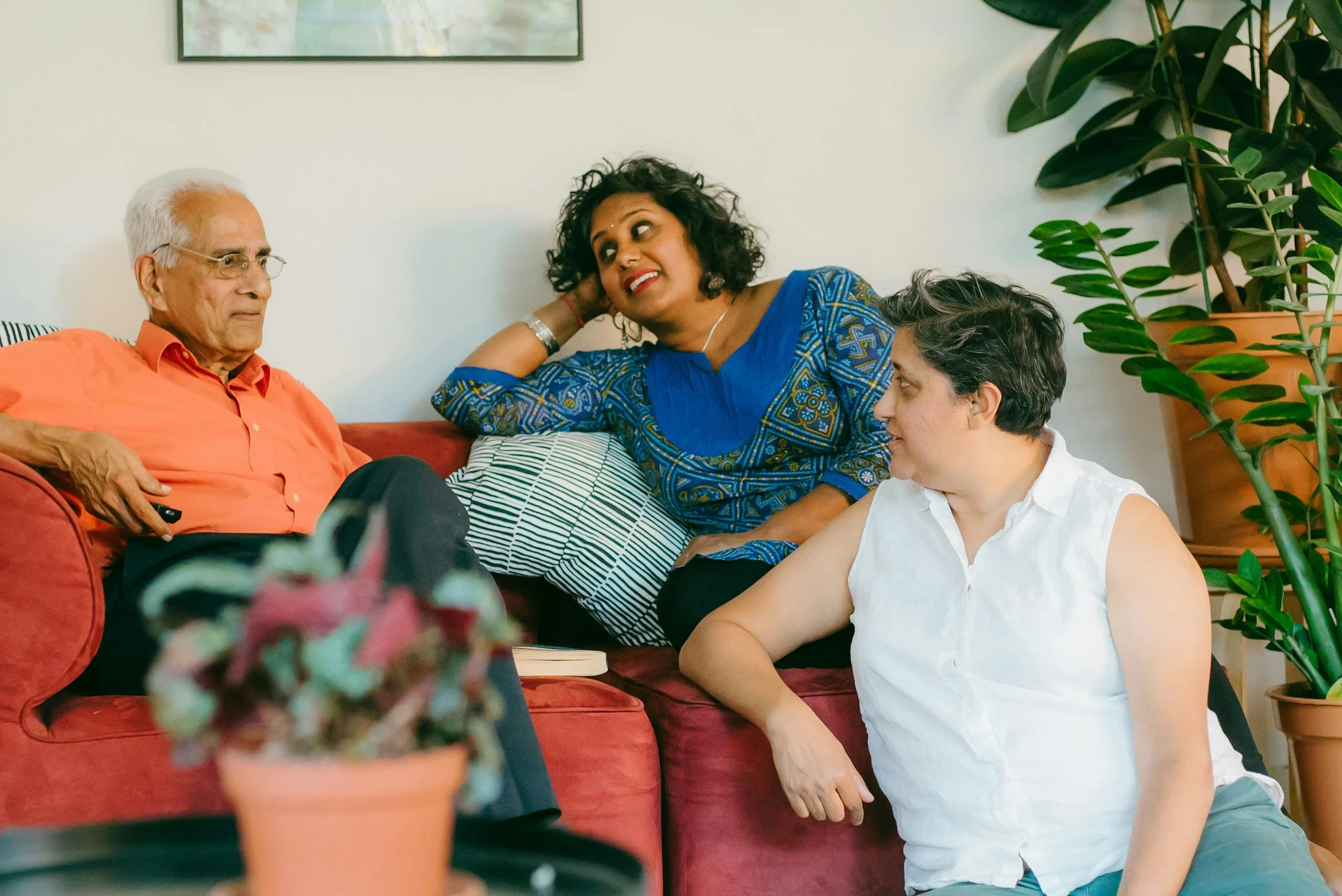 Three people engaged in a conversation in a living room. An elderly man wearing glasses and an orange shirt sits on a red couch, a woman with curly dark hair and a blue patterned dress leans against the wall, and another woman with short dark hair and a white sleeveless shirt kneels on the floor, all surrounded by potted greenery.