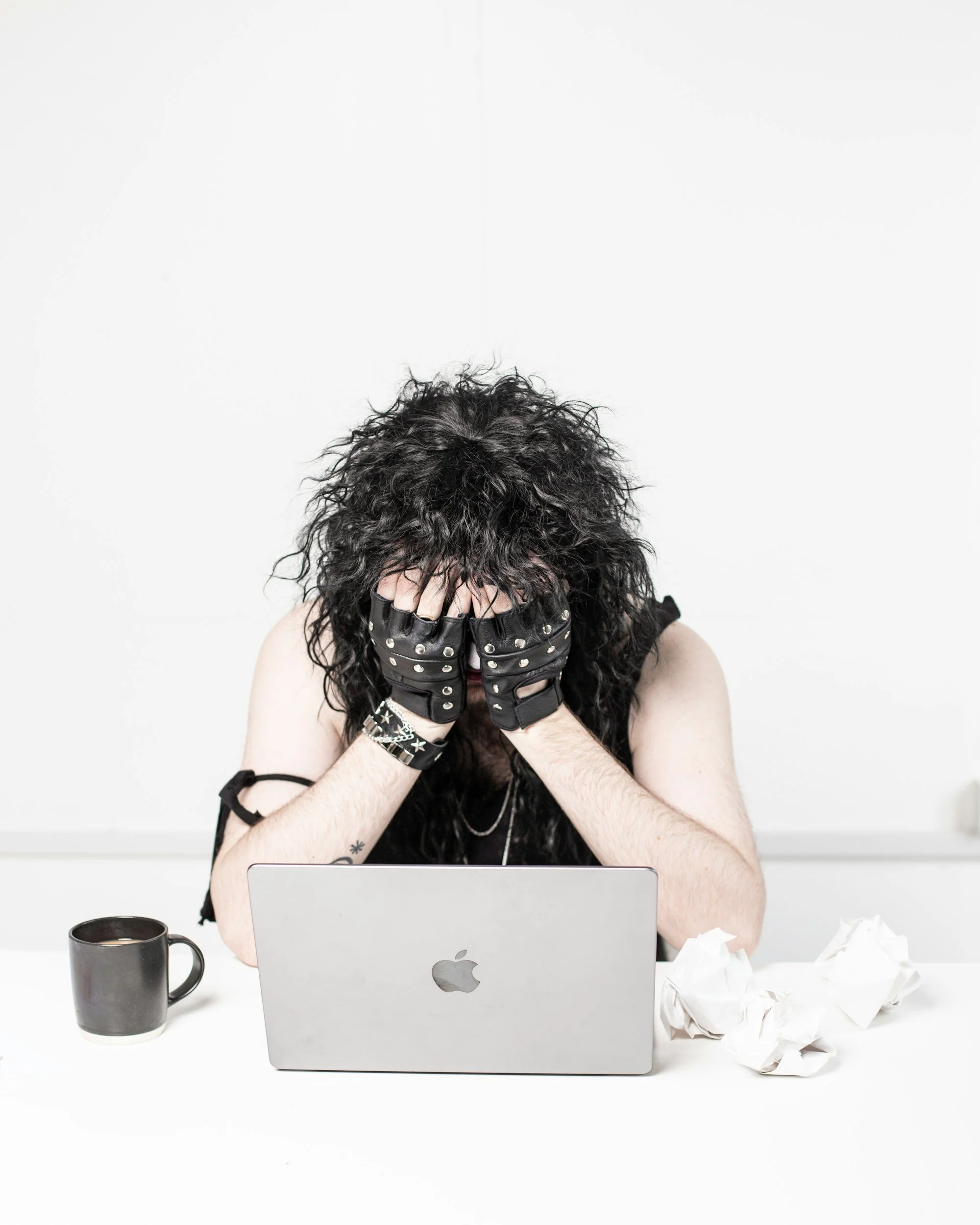 Person with curly black hair covering face with hands, wearing black gloves with metal studs, sitting at a white desk with a silver MacBook laptop, a black coffee mug, crumpled tissue papers, and a white wall in the background.