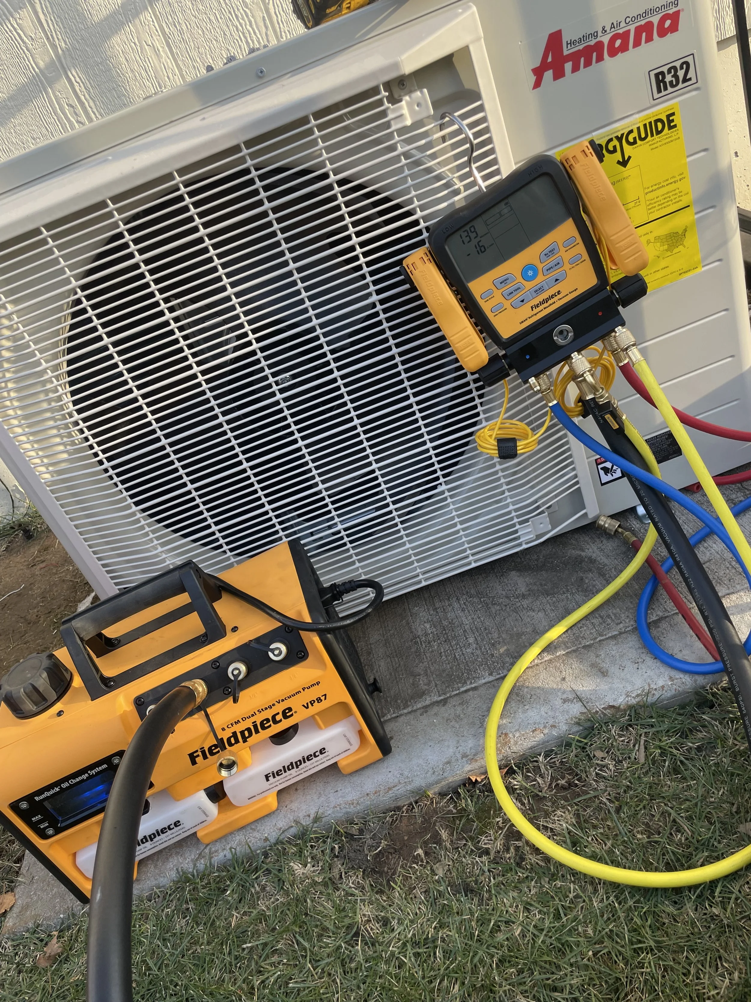 An outdoor communicating mini split heat pump unit and a yellow vacuum pump connected by hoses, with a digital manifold gauge placed on the HVAC unit for refrigeration or air conditioning system diagnostics.