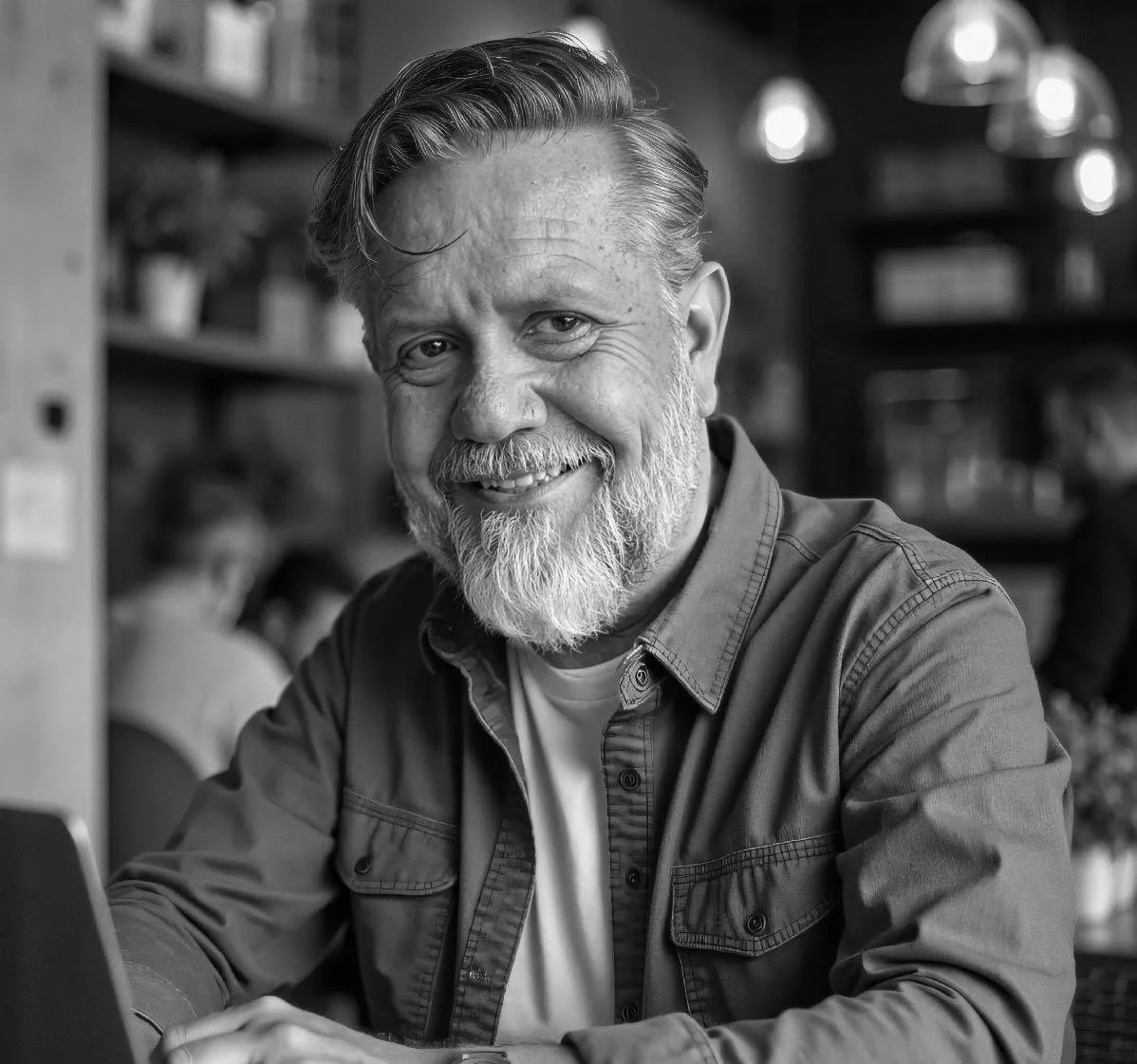 Ron Maxson Jr, founder of The Dads Prayer Coach, smiling; a man with a gray beard and mustache, wearing a casual button-up shirt, sitting at a table in a cozy indoor setting, possibly a cafe or library, with blurred background shelves and lights.