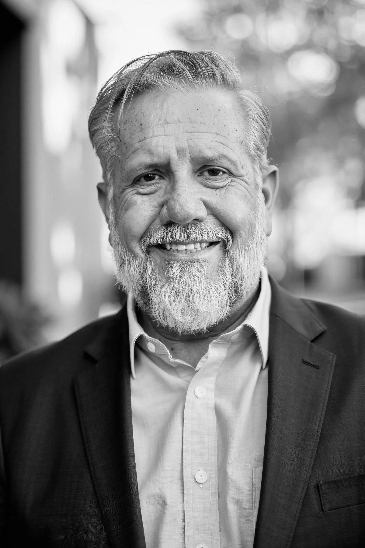 Monochrome image of Ron Maxson Jr standing outdoors in nature with open sport coat and dress shirt, smiling. The Dads Prayer Coach.