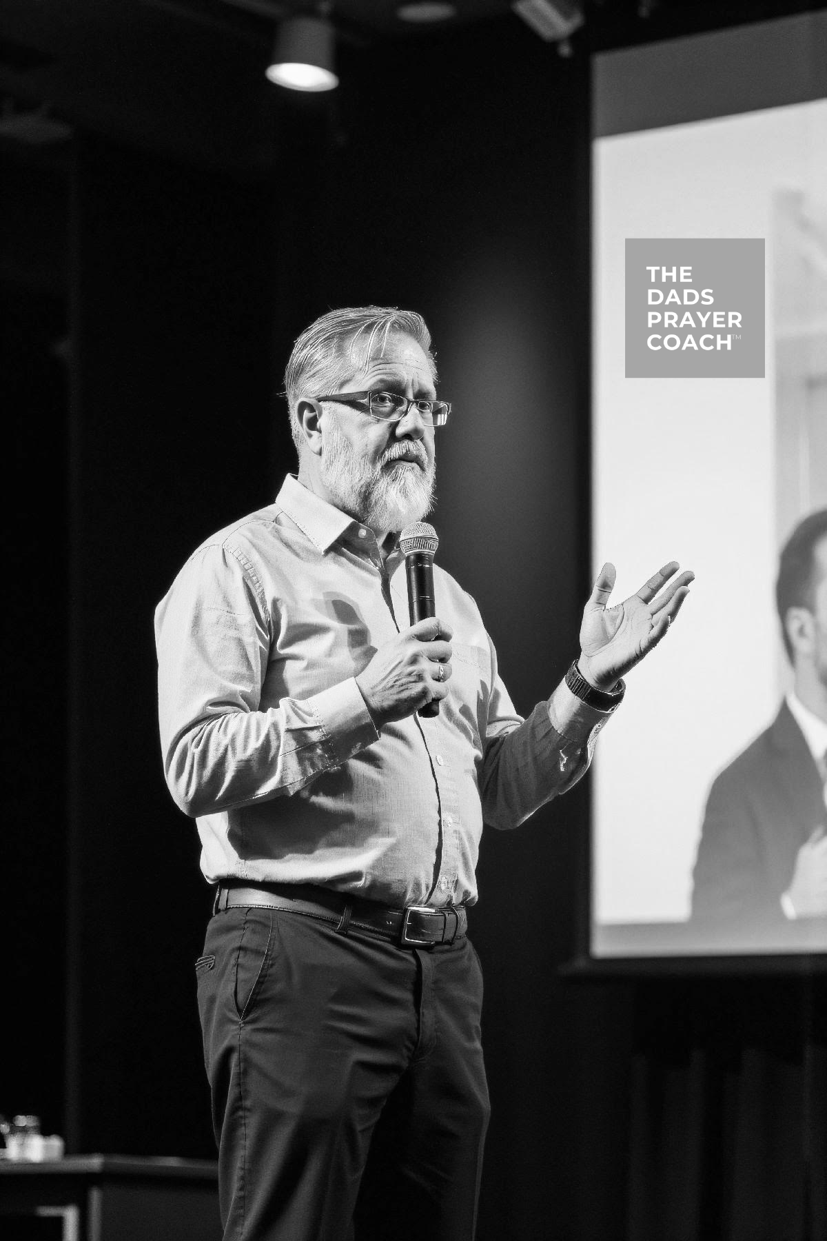 Ron Maxson Jr standing on stage with microphone in hand pointing to a video screen and speaking to a group of men.