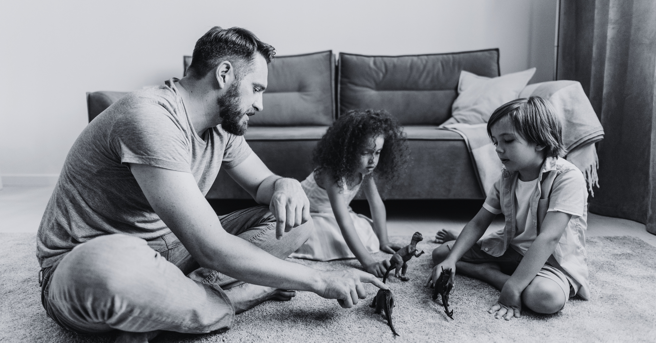A man and two children sitting on a carpeted floor, playing with toy dinosaurs in a living room with a sofa and pillows in the background.