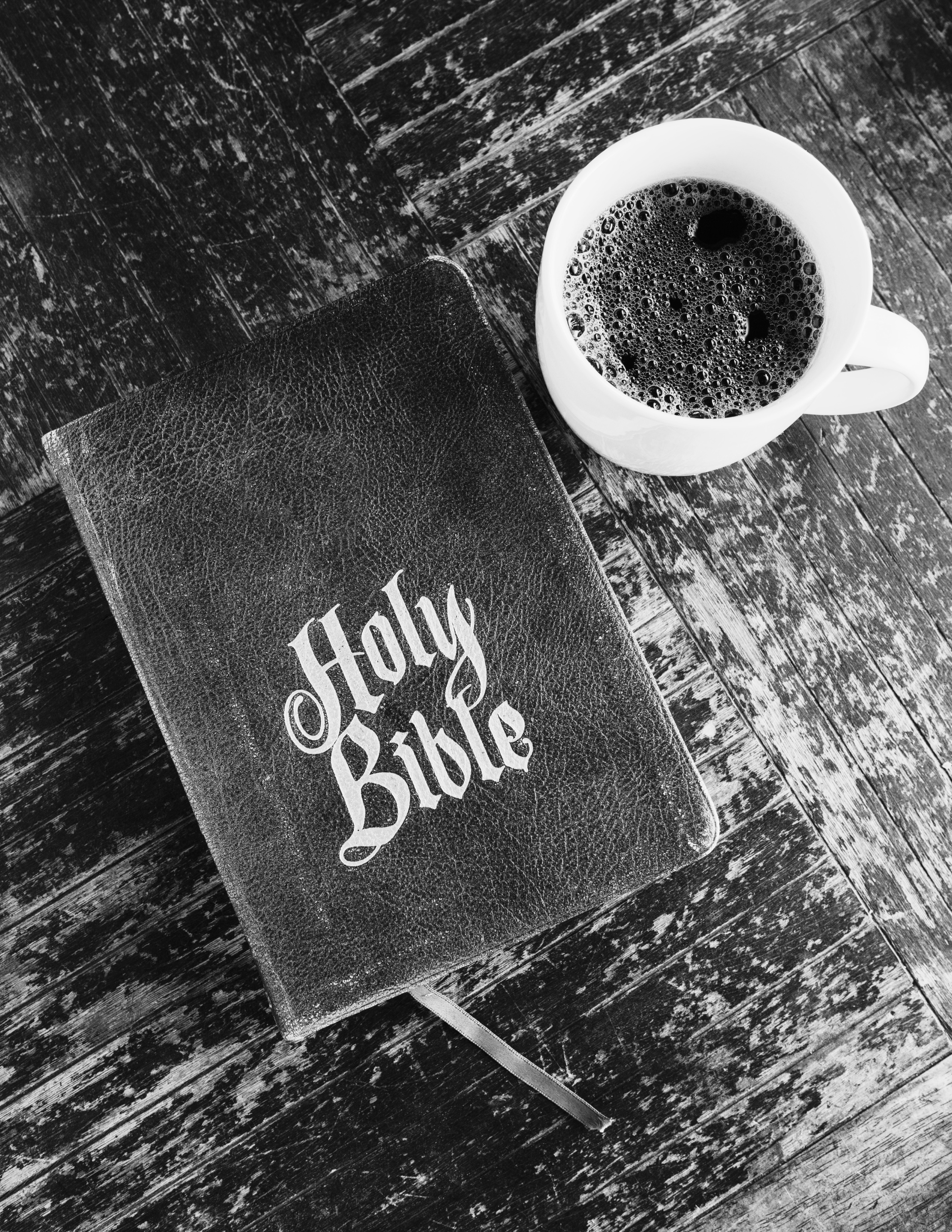 A black and white image showing a book titled "Holy Bible" with a textured cover, placed on a worn wooden surface. Next to the book, there is a white coffee mug filled with black coffee, with foam bubbles visible on the surface.