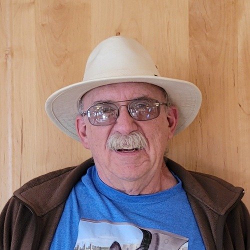 An elderly man with glasses and a mustache, wearing a white hat and a blue shirt, sitting in front of a wooden background.