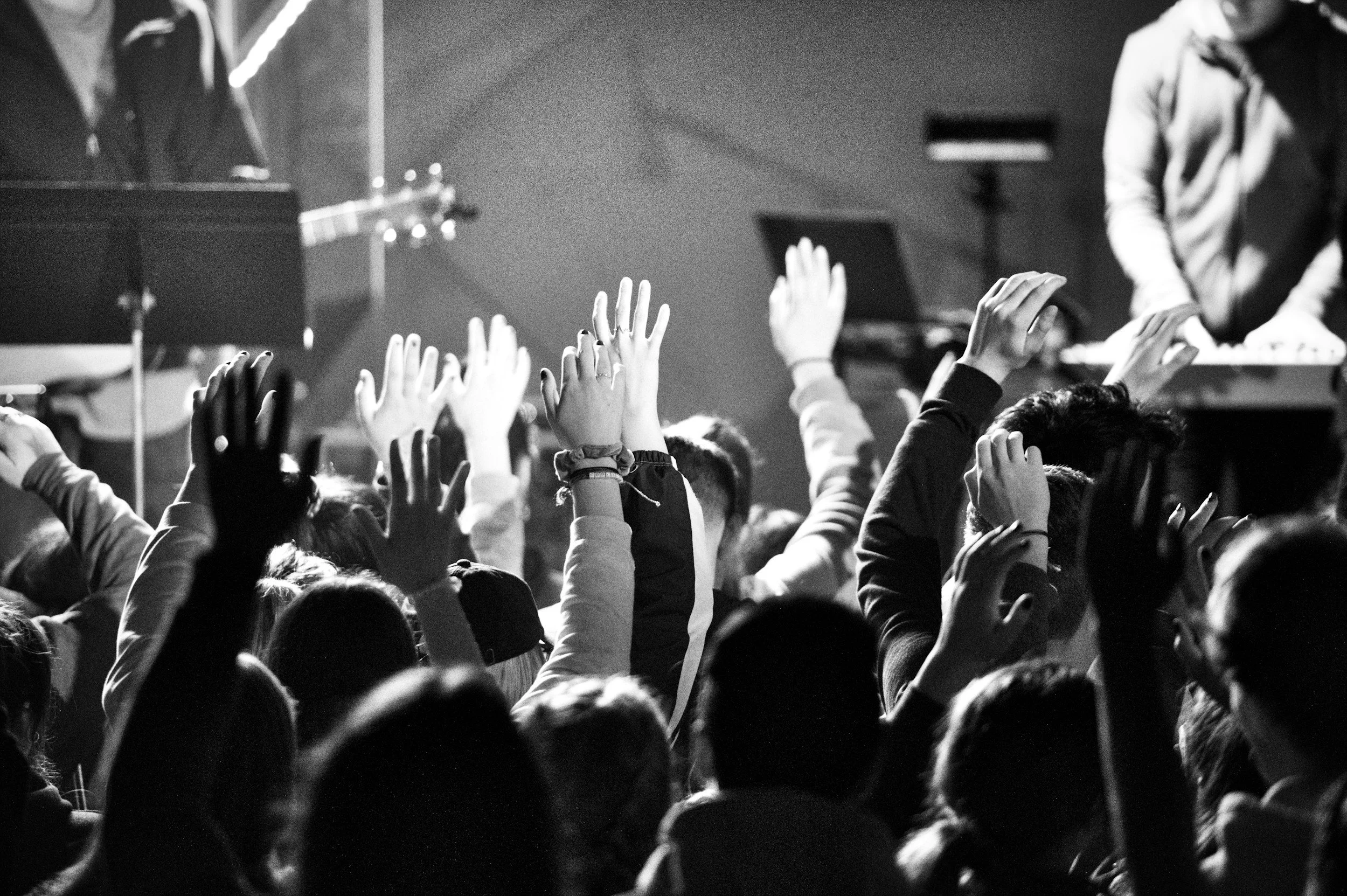 Black and white photo of a crowd with their hands raised at a concert or event, with some people on stage or near a DJ in the background.