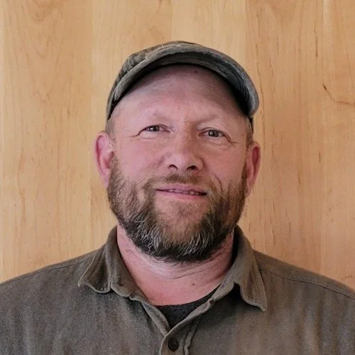 A man with a beard wearing a baseball cap and a brown shirt, standing in front of a wooden wall. Jason Lyon, Elder of Dalkena Community Church.