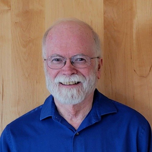 Smiling elderly man with glasses and a white beard, wearing a blue shirt, standing in front of a wood-paneled wall. Steve Wilson, Elder of Dalkena Community Church.