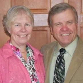 A smiling older woman with short blonde hair wearing a pink jacket and floral blouse, standing next to a smiling middle-aged man with short gray hair wearing a beige suit and striped tie, in front of wooden paneling.