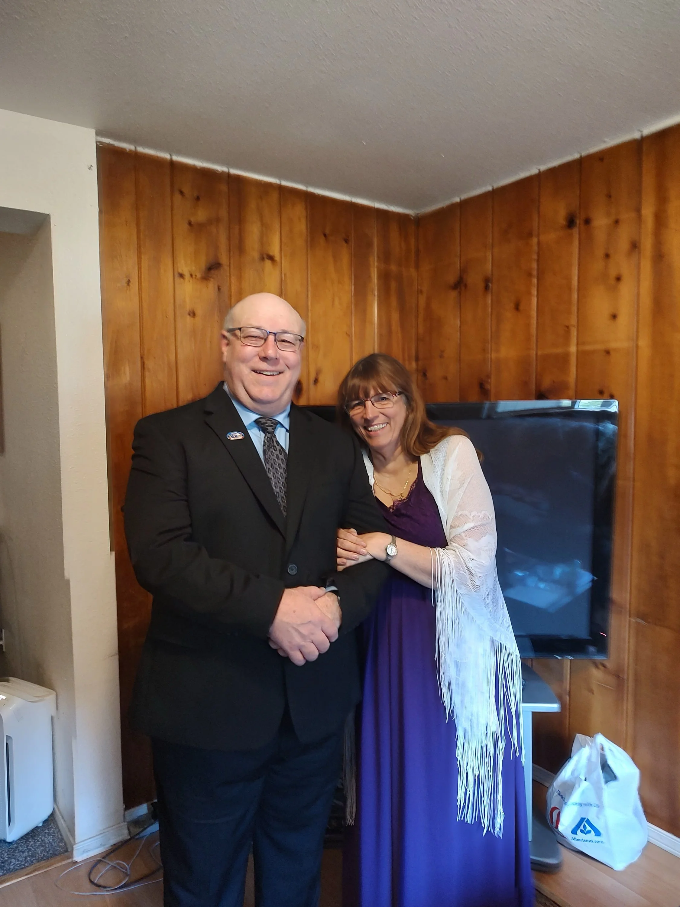 A man in a suit shaking hands with a woman in a purple dress and white shawl, both smiling, standing in a room with wood paneled walls. Anita Alford and Ken Alford.